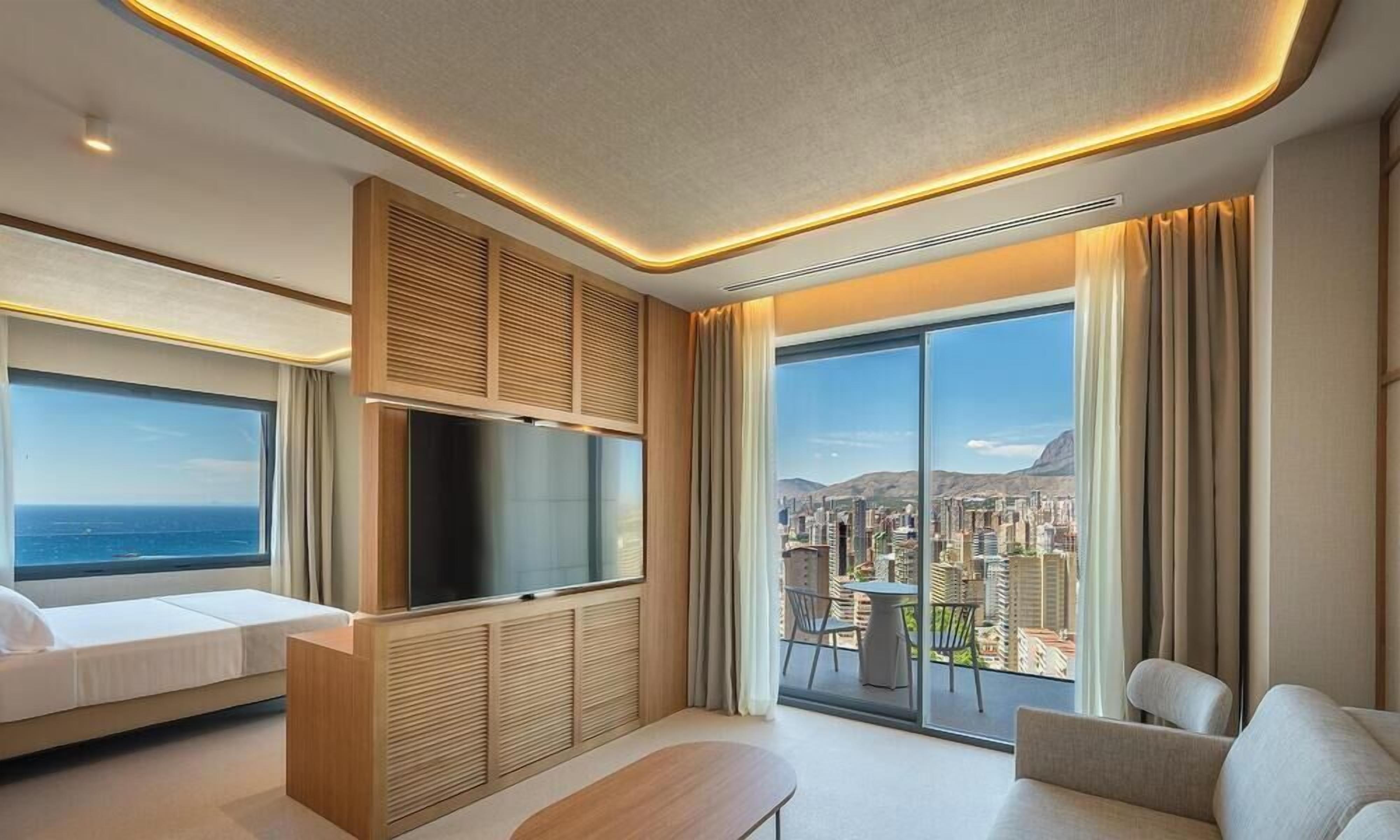 Habitación de hotel moderna con un ventanal grande que ofrece vistas de la ciudad, un televisor de pantalla plana, un sofá y una mesa de comedor.
