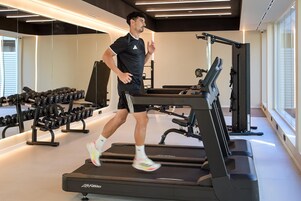 A person running on a treadmill in a gym with weightlifting equipment and dumbbells.