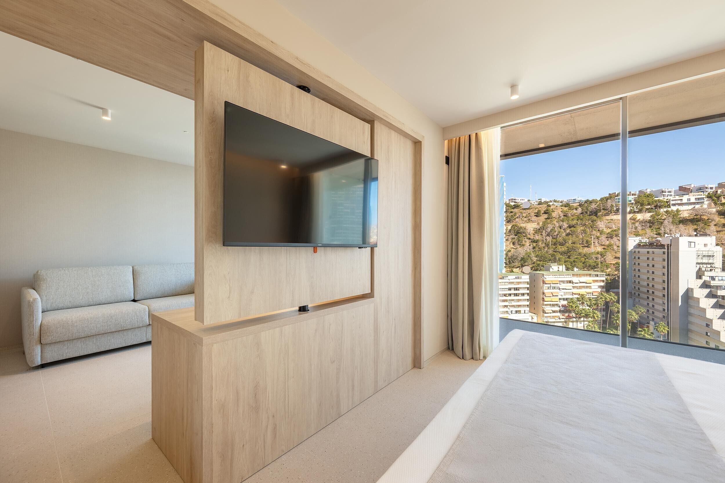 Habitación de hotel moderna con un televisor grande montado en una pared de madera, un sofá gris y un balcón con vistas a la ciudad.