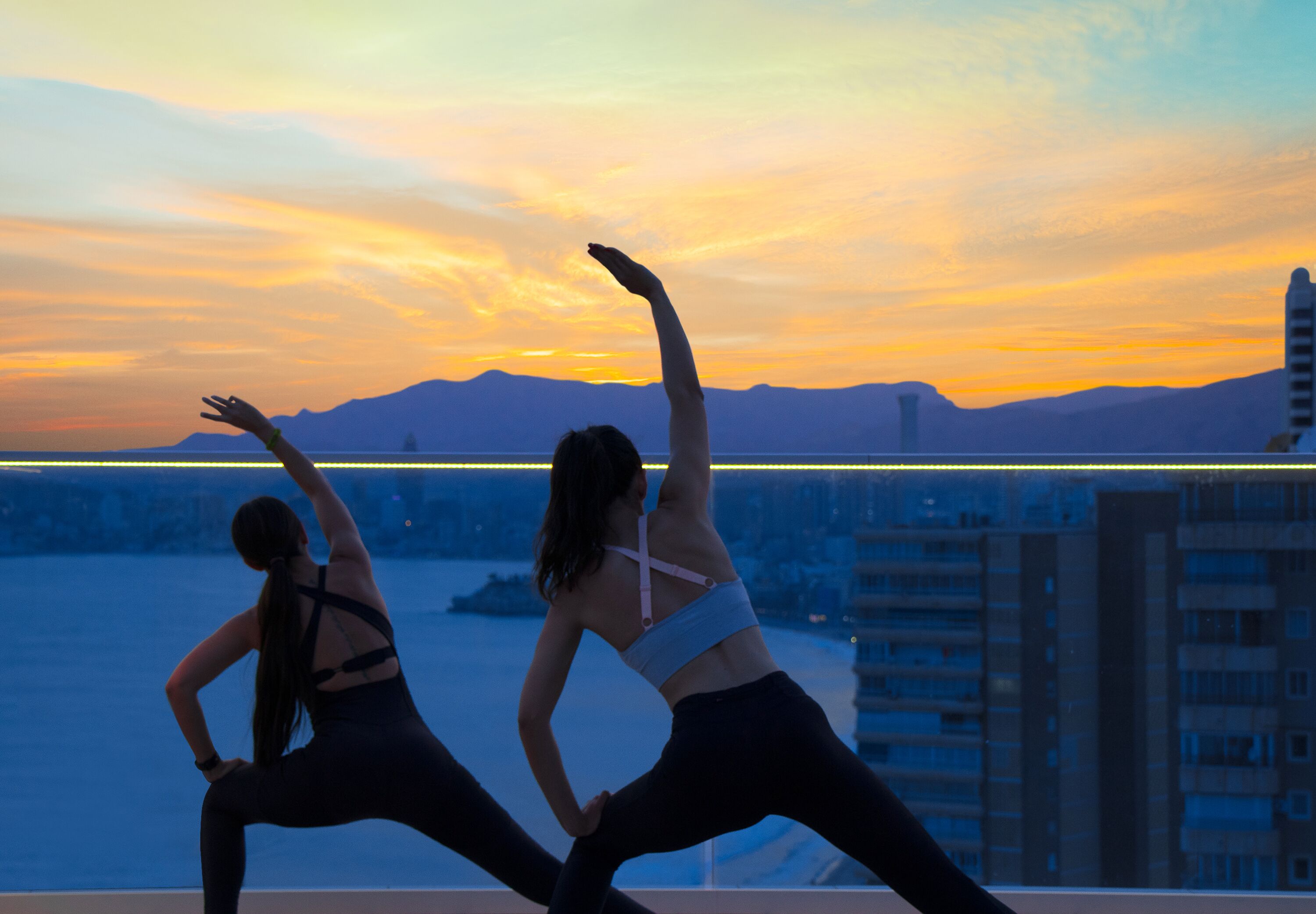 Two people stretching on a balcony with a cityscape and mountains in the background.