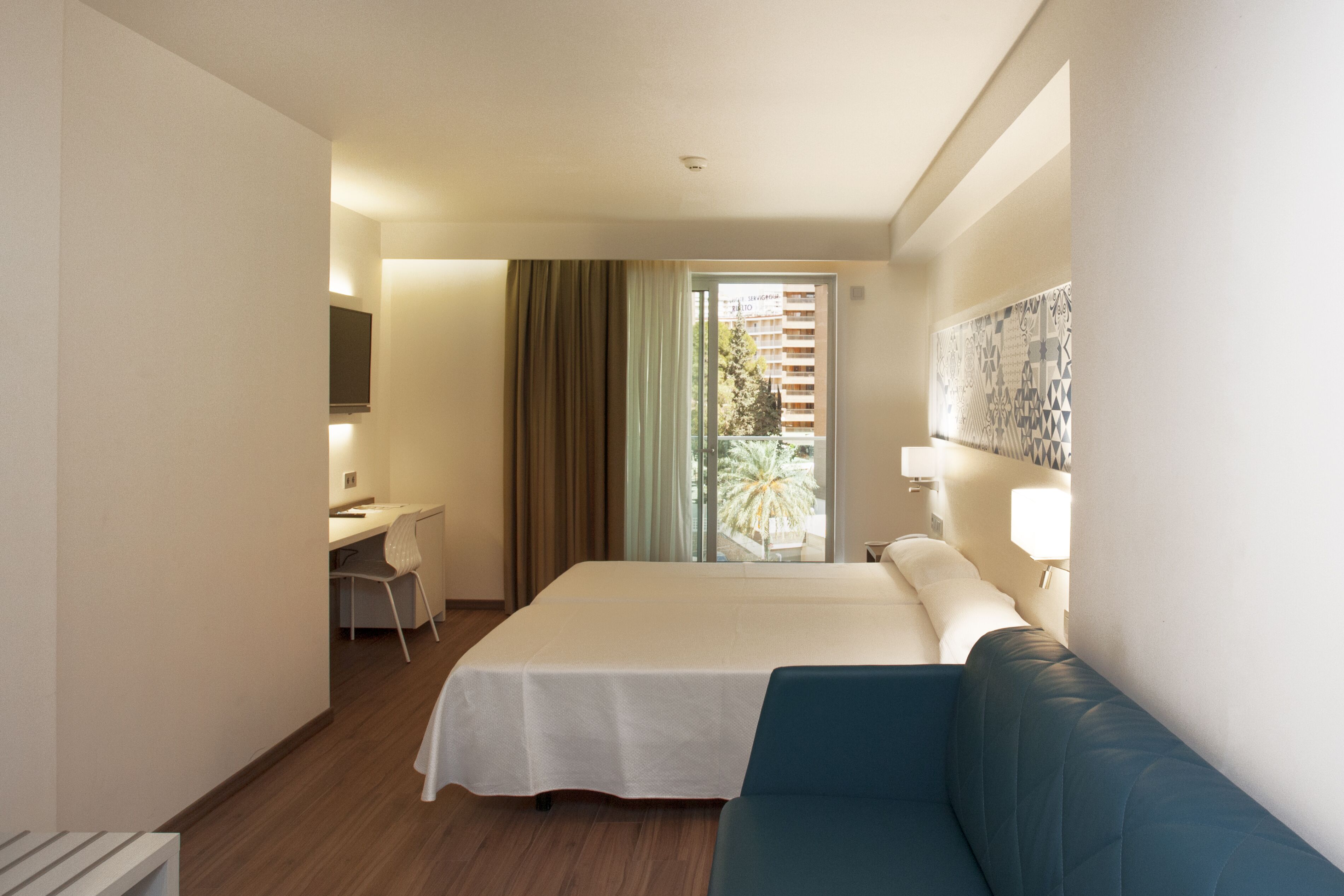 A hotel room with a bed, a desk, a chair, a TV, a window with curtains, and a view of buildings and trees.