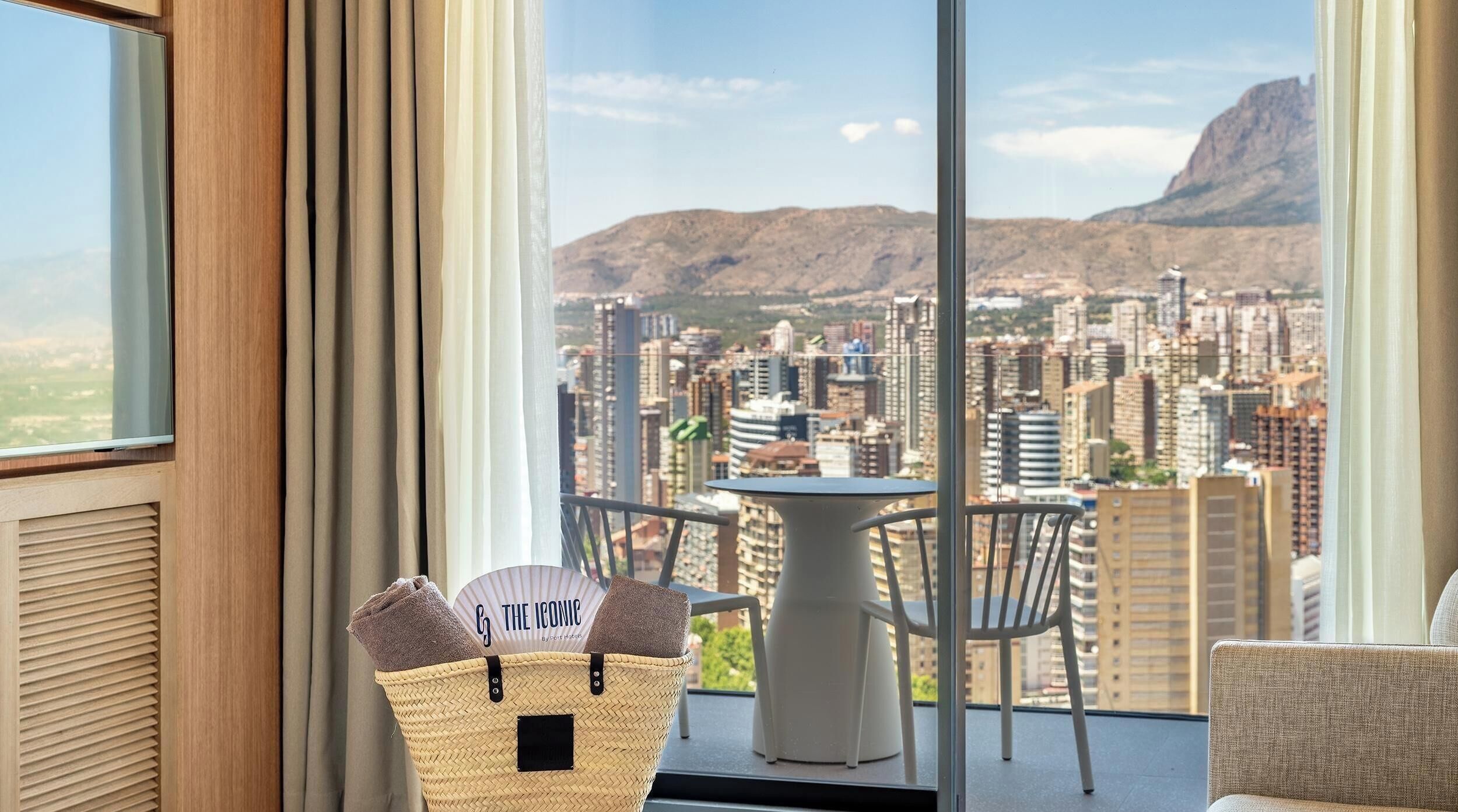 A balcony with a view of a cityscape and mountains, a table with chairs, and a basket with towels.