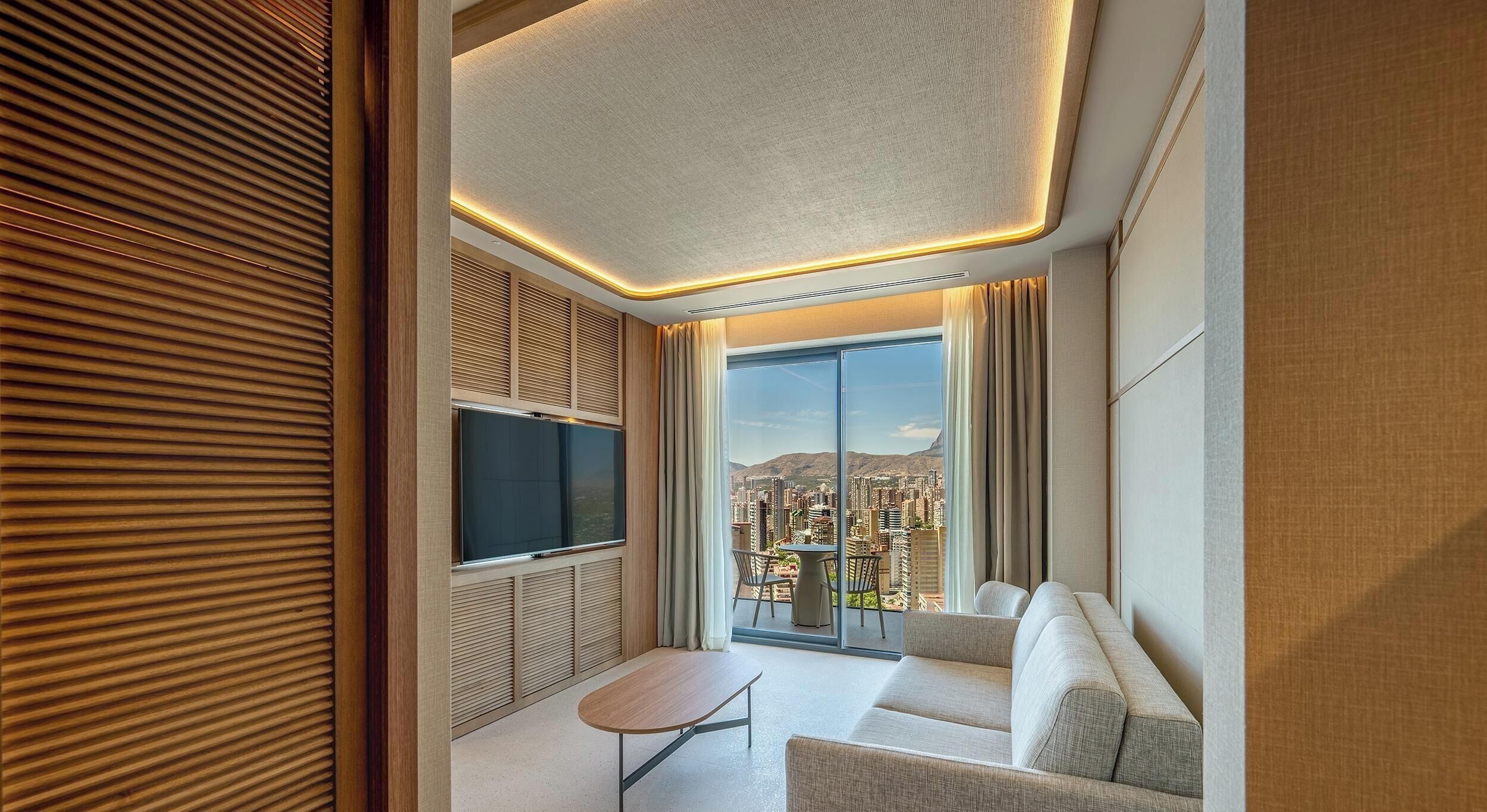 Una habitación de hotel moderna con un ventanal grande, un televisor de pantalla plana, un sofá y una mesita.