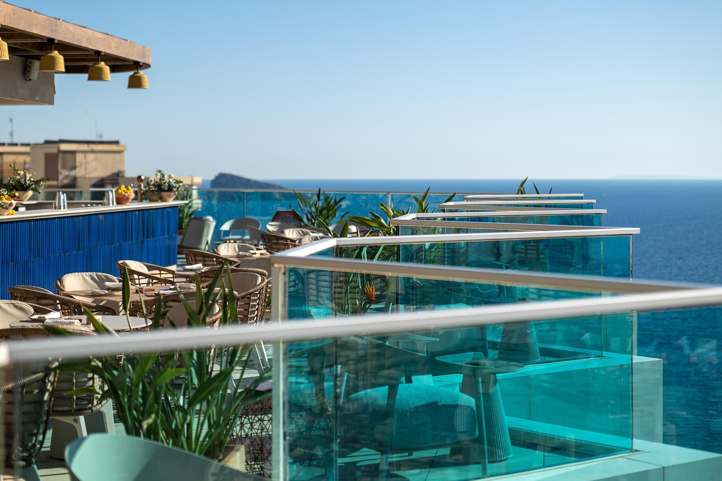 A rooftop terrace with glass balustrades overlooking the sea, furnished with lounge chairs and tables.