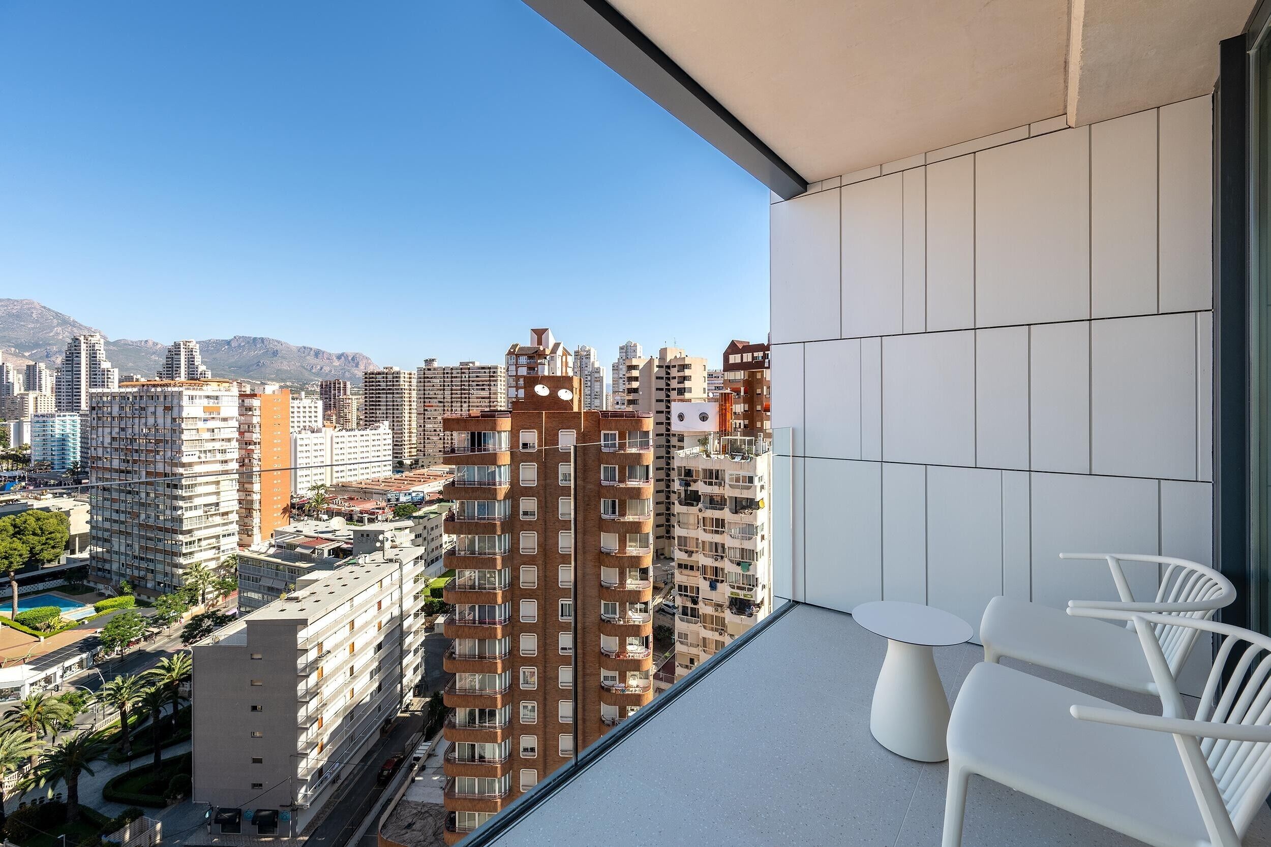 A balcony with white chairs and a view of a cityscape with tall buildings and mountains in the background.