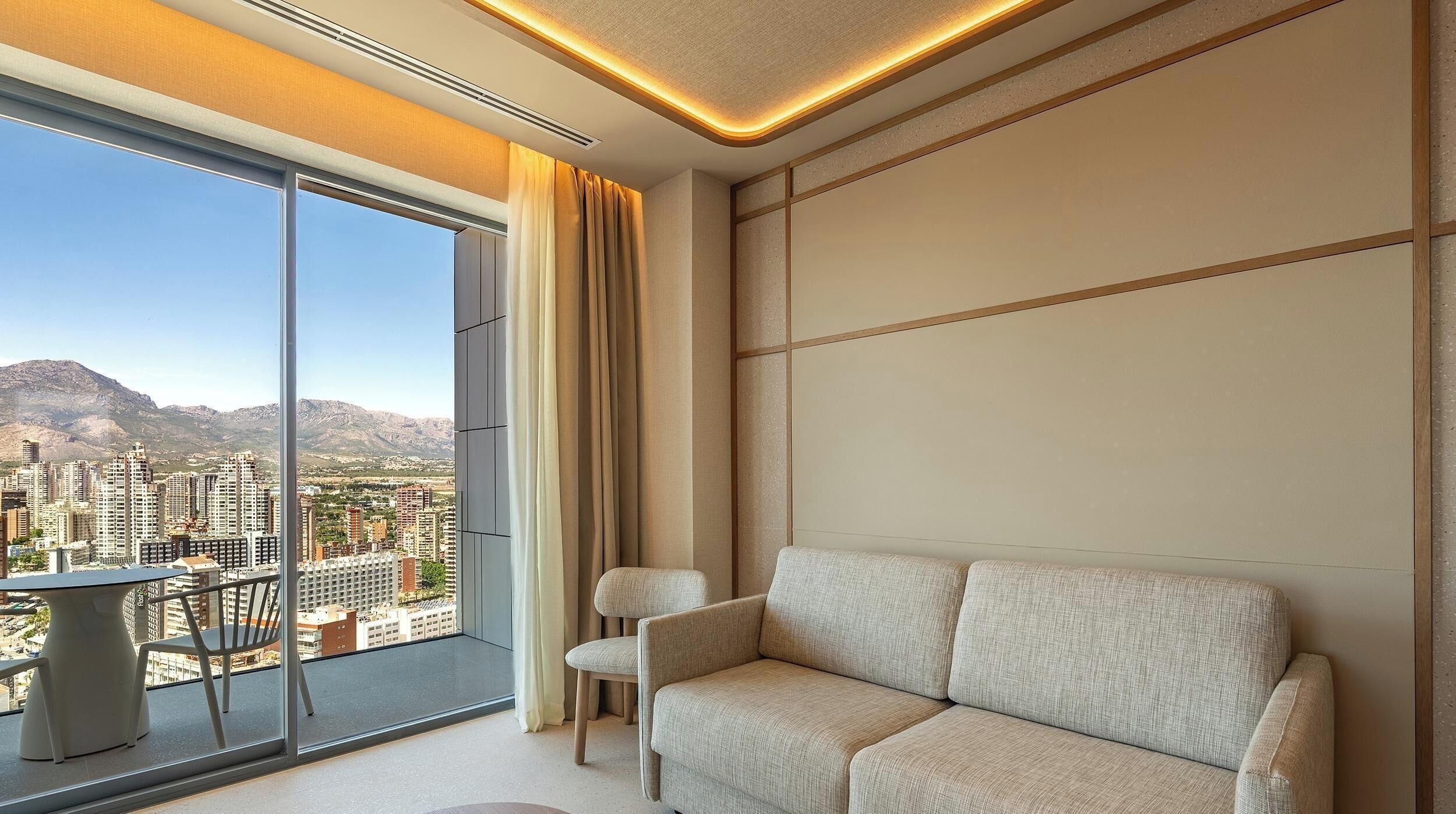 A modern living room with a sofa, a chair, and a table. There is a large window offering a city view.