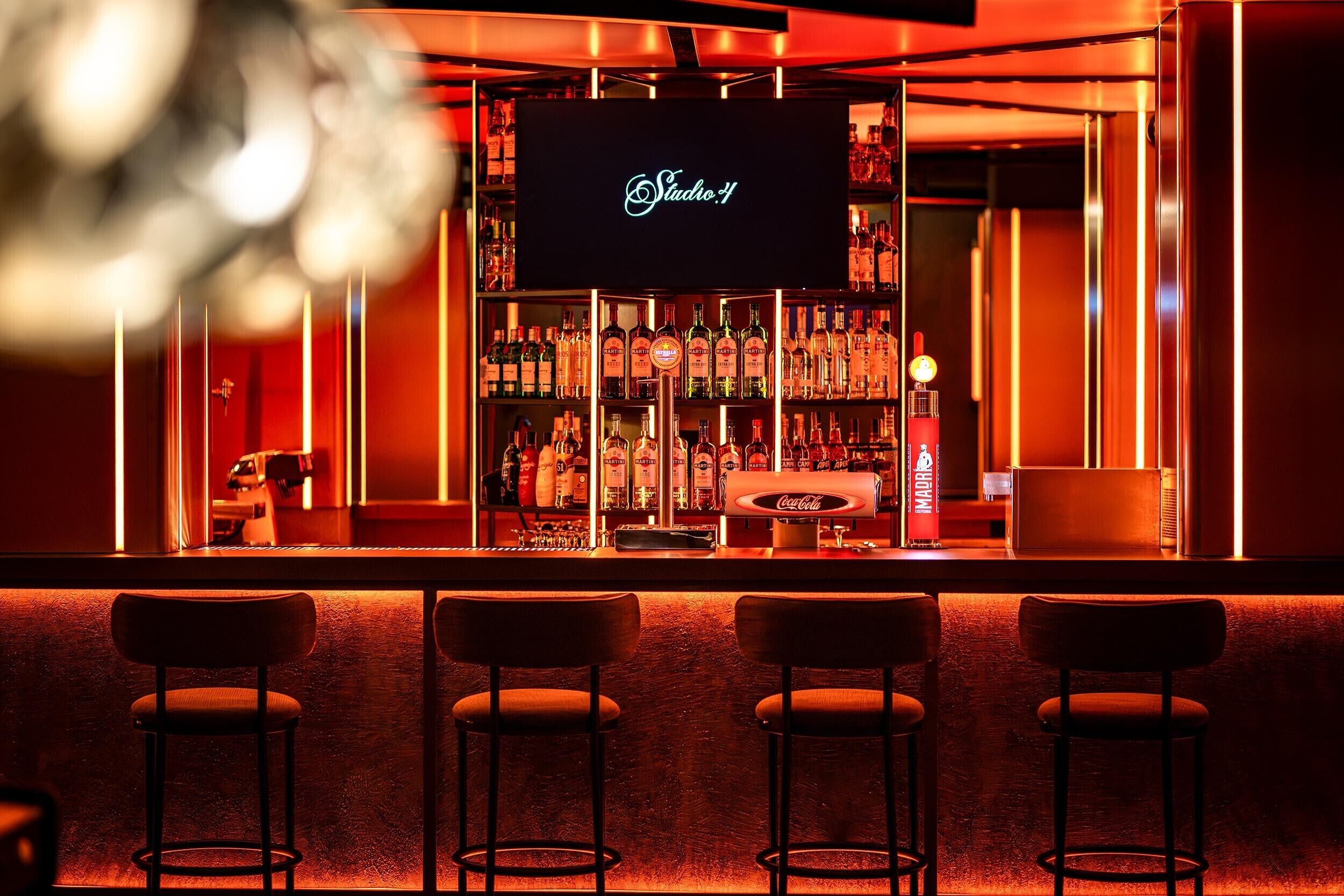 Zona de bar con un letrero de neón que dice 'Studio 4', estantes llenos de licores y tres taburetes de bar.