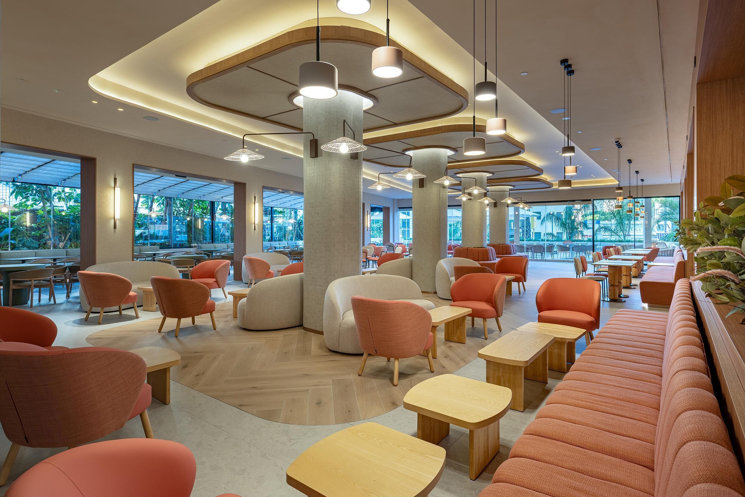 A modern indoor space with seating areas, tables, and large windows offering views of greenery.
