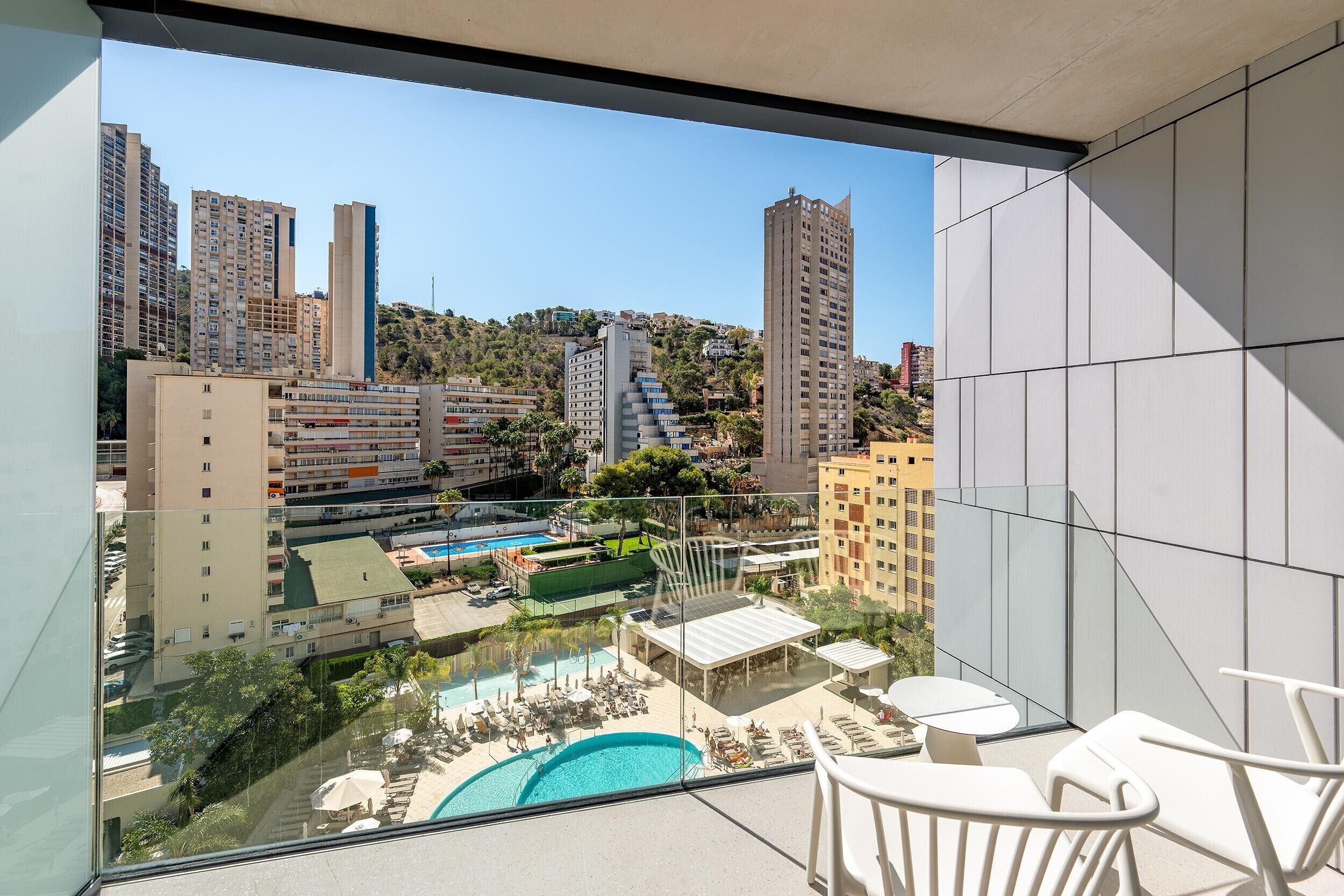 A balcony with a view of a swimming pool, lounge chairs, and a cityscape with tall buildings.