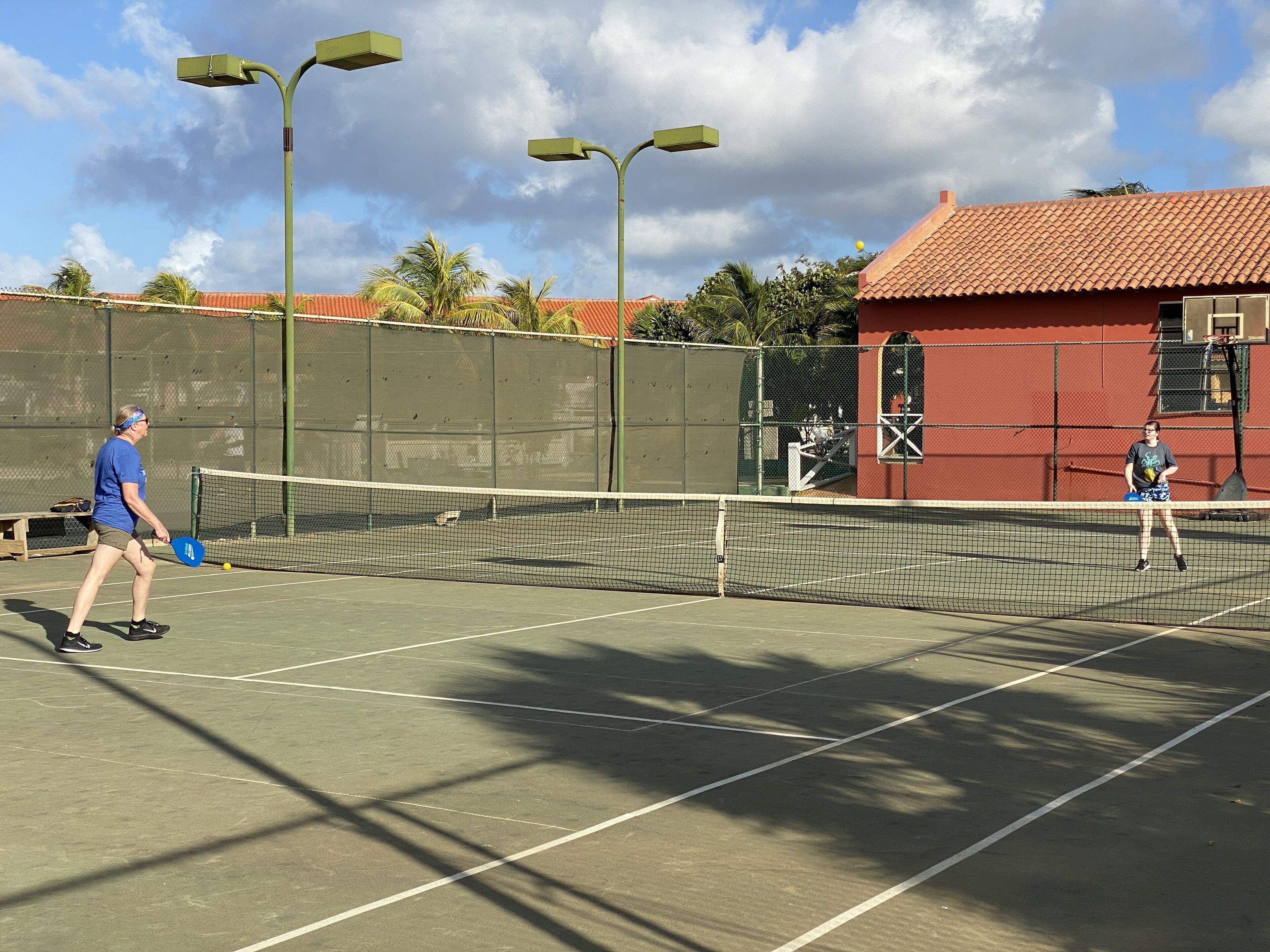 An outdoor tennis court with a net, two players, and a red building in the background.