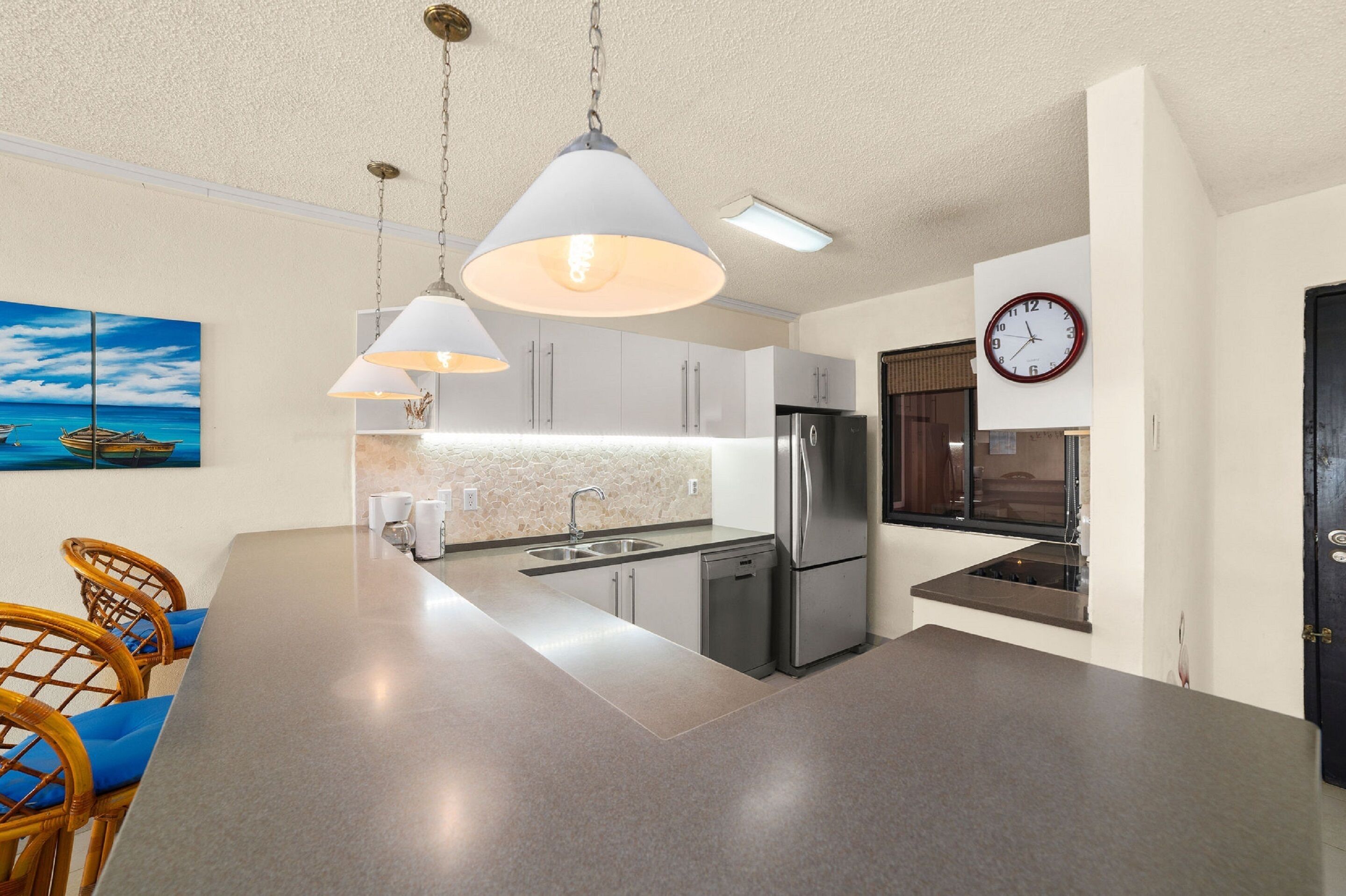 Uma cozinha moderna com uma ilha grande, eletrodomésticos de aço inoxidável, iluminação pendente e um relógio na parede.