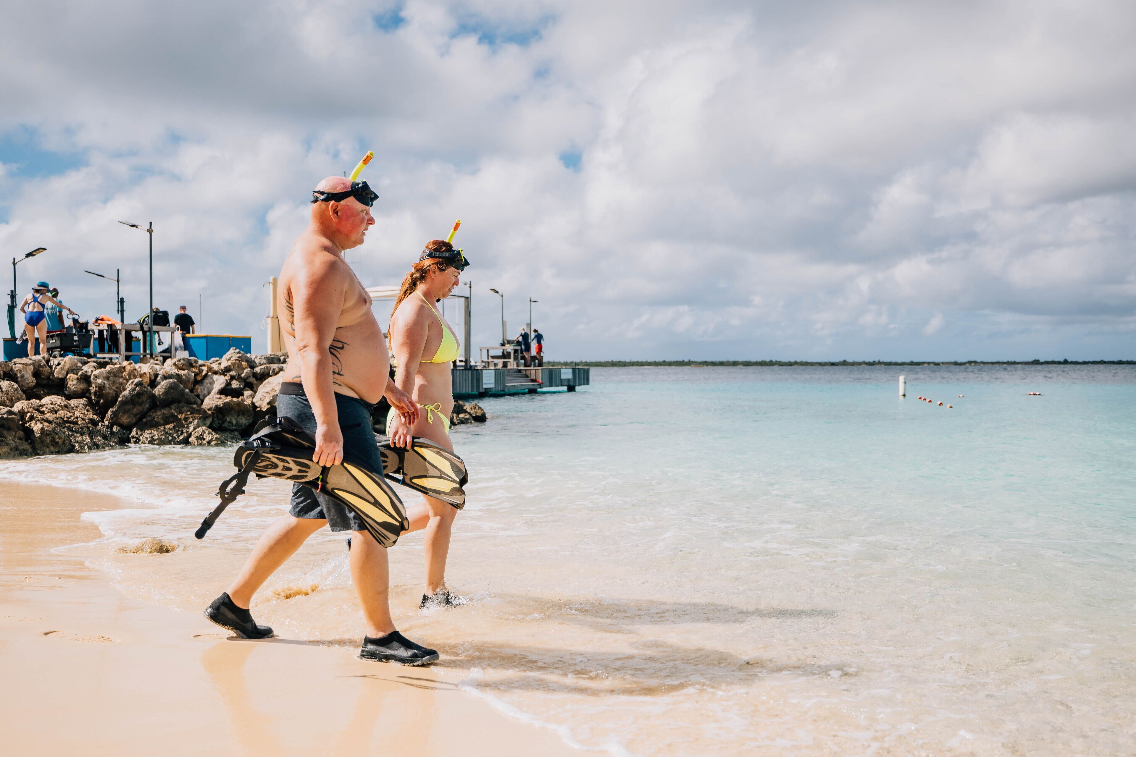 Une scène de plage où l’on voit un homme et une femme se promener le long du rivage, tenant du matériel de plongée avec tuba.