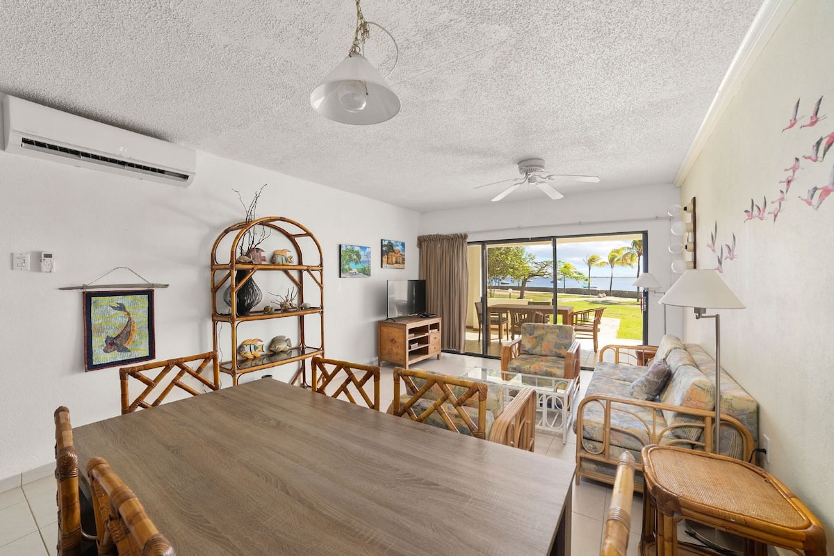 A spacious living room with a wooden dining table, wicker furniture, a ceiling fan, and a view of the outdoors.