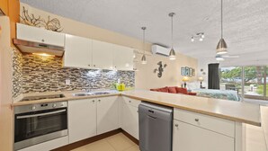 A modern kitchen with white cabinets, a tiled backsplash, and a dishwasher. There is a dining area with a table and chairs visible through a large window. - Sand Dollar Bonaire (Kralendijk)