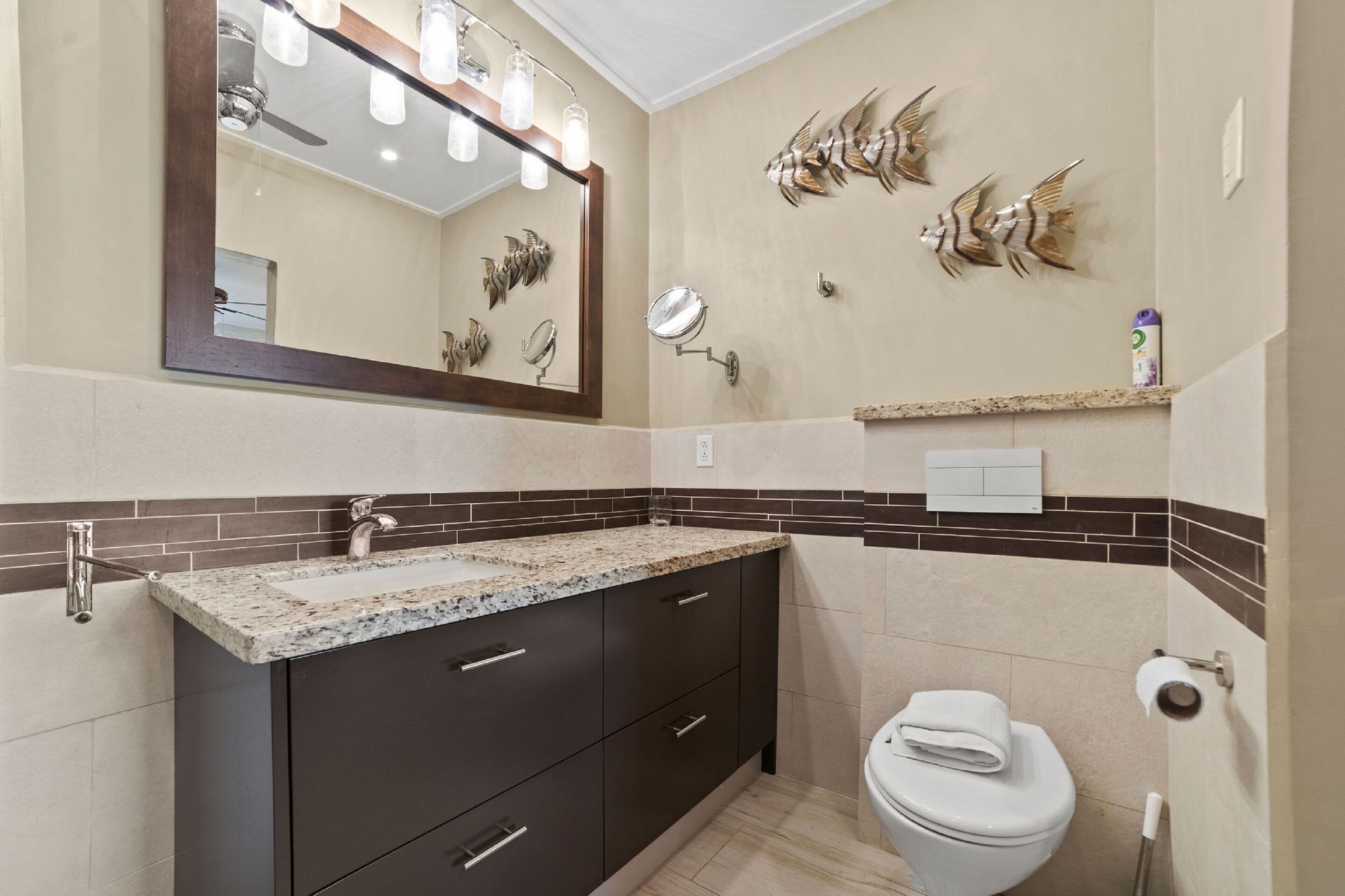 Une salle de bain dotée d’un comptoir en granit, de meubles sombres, de toilettes blanches et d’œuvres d’art décoratives sur les murs.