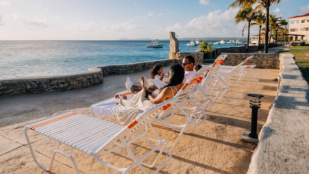 A beachside lounge with white sun loungers, a stone wall, and a view of the sea with boats. - Sand Dollar Bonaire (Kralendijk)