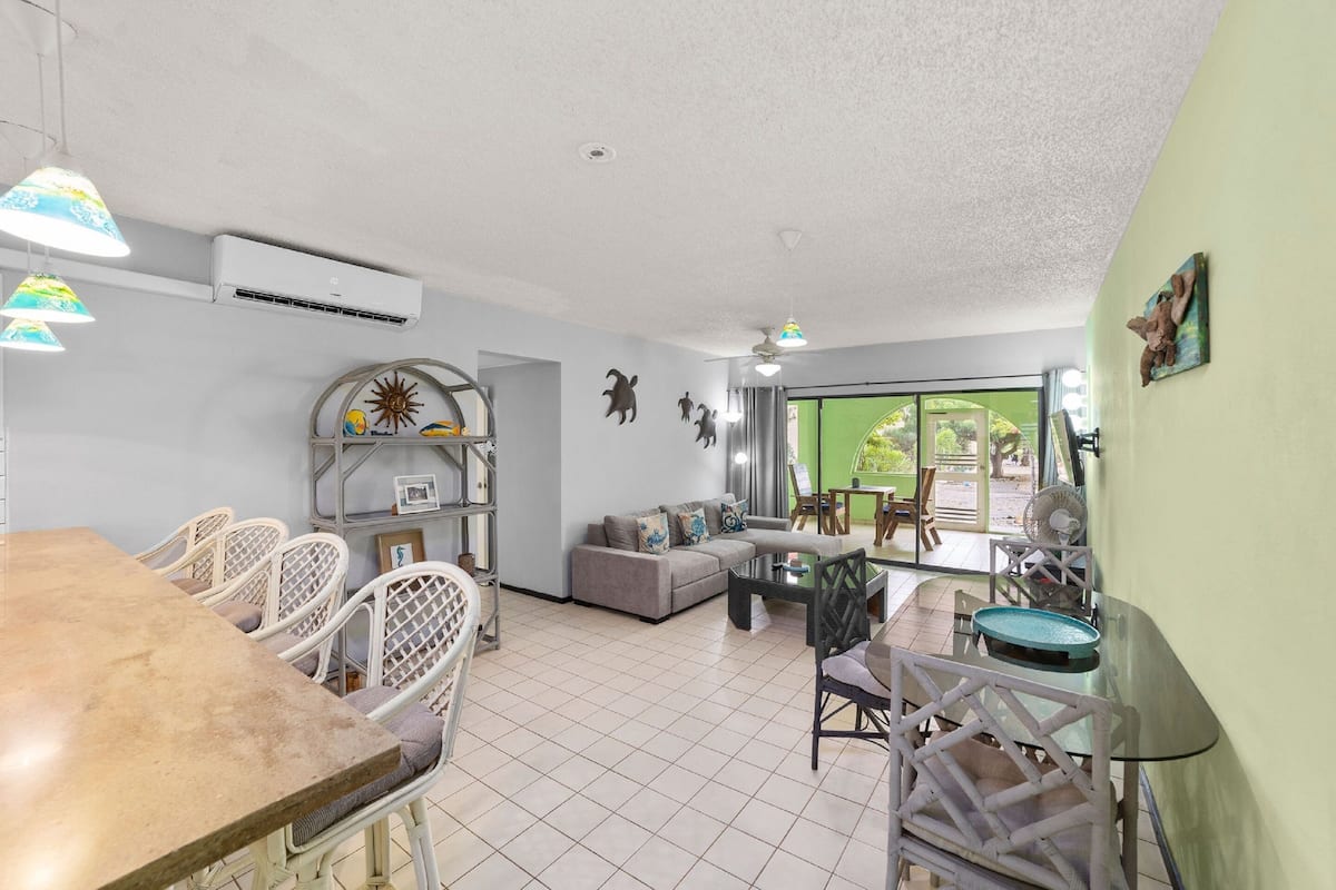 A spacious living area with a dining table, chairs, a sofa, and a glass coffee table. There is a large window providing a view of the outdoors.