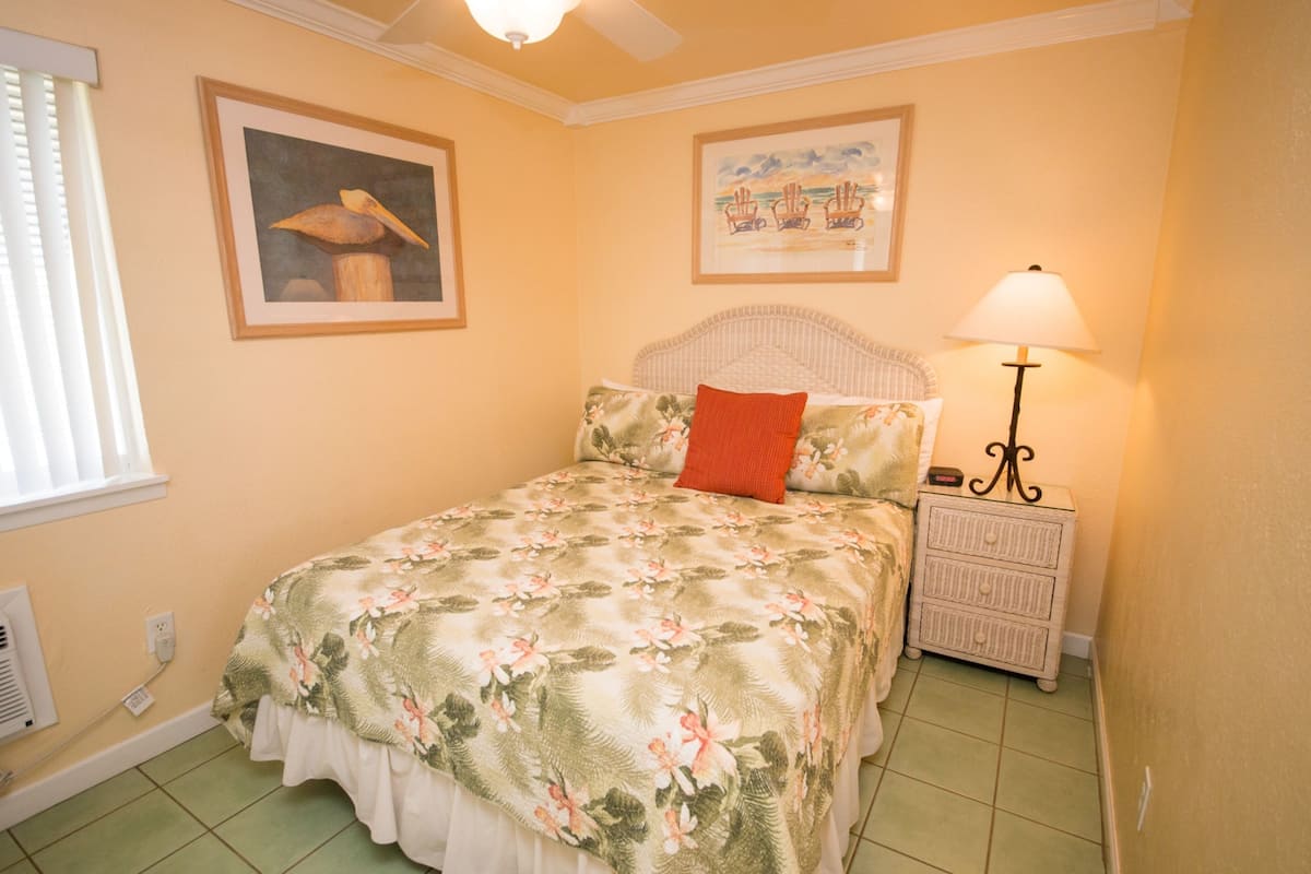 Un dormitorio con colcha estampada, almohada roja, mesita de noche con lámpara y dos cuadros en la pared.