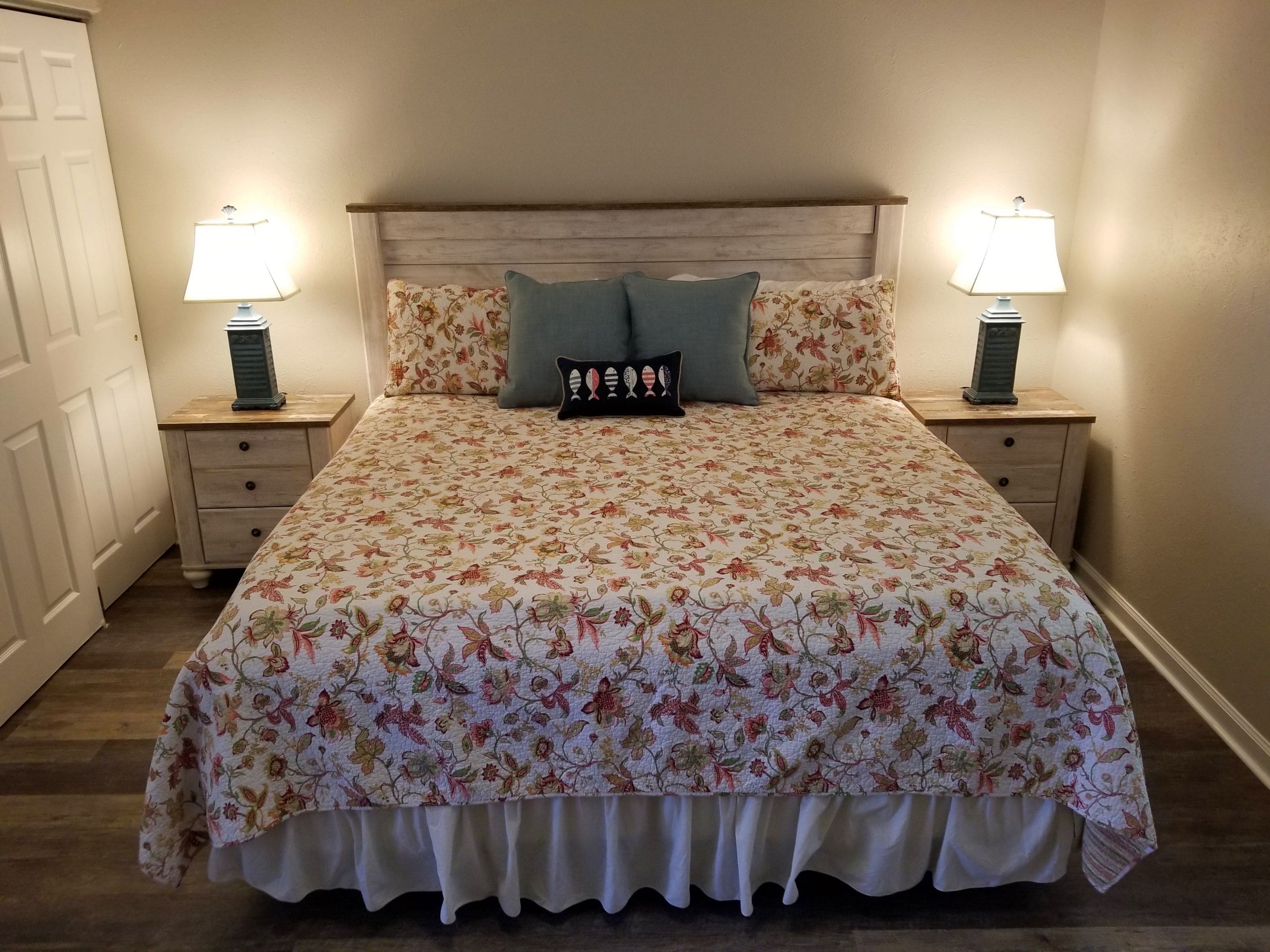 A bedroom with a floral bedspread, two nightstands with lamps, a wooden headboard, and a door.