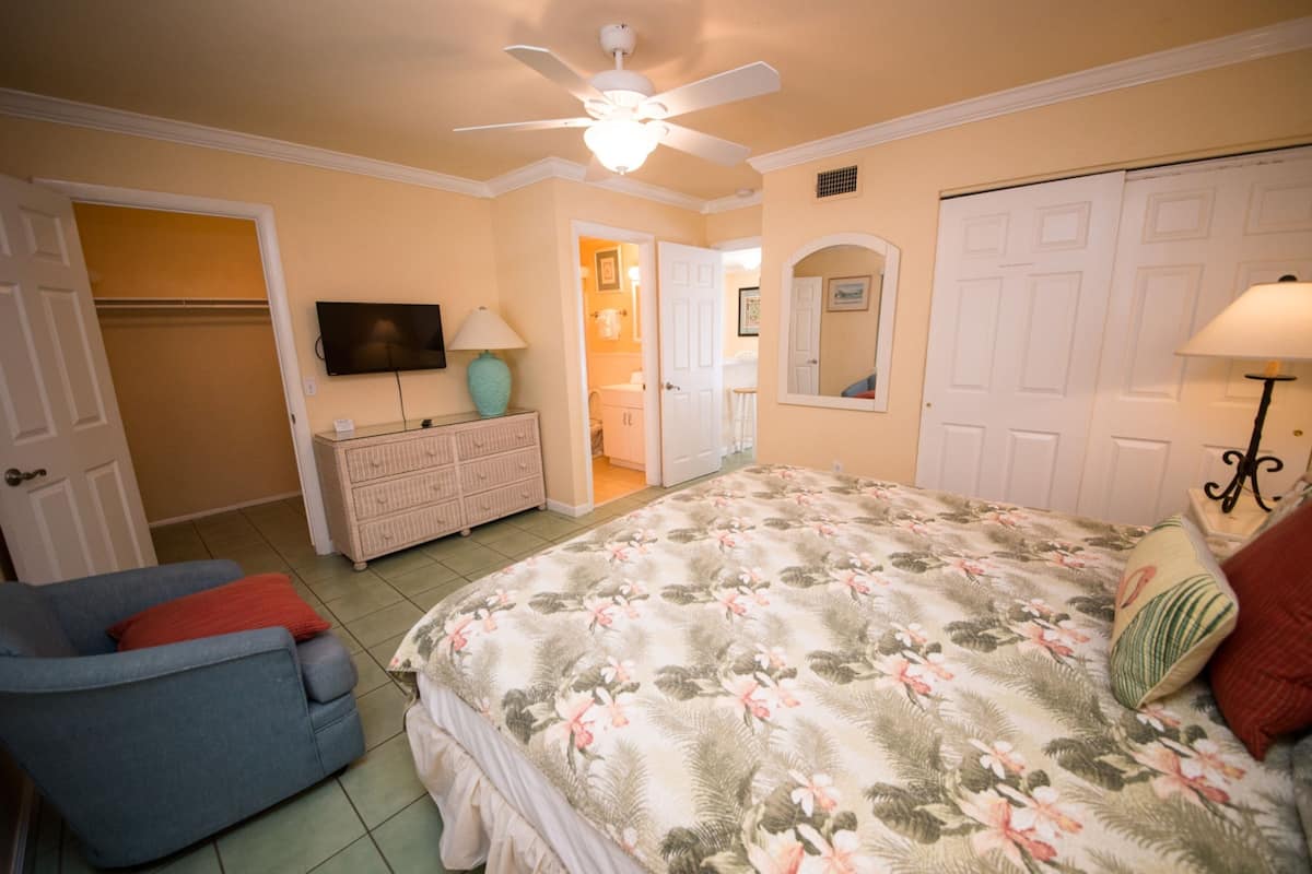 Un dormitorio con cama, una silla, cómoda, televisor y ventilador de techo.