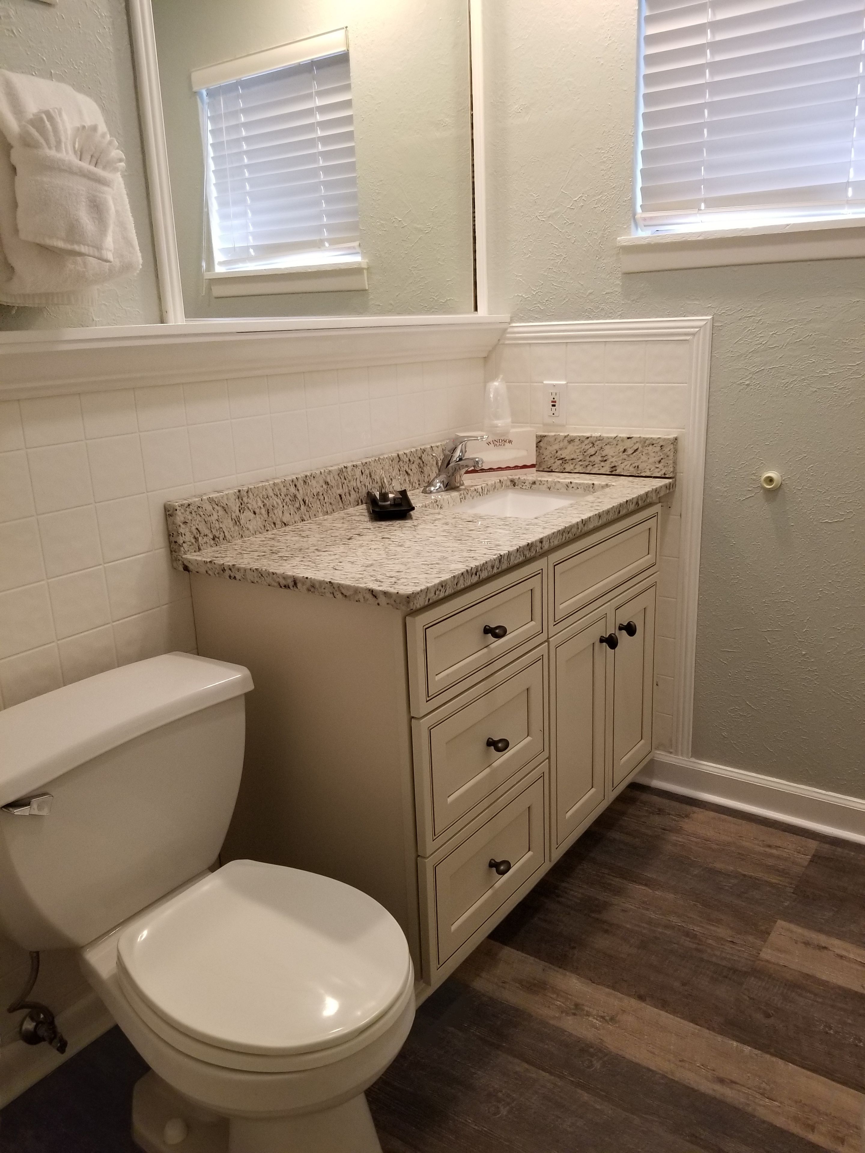 Une salle de bain avec des toilettes blanches, un lavabo surmonté d’un plan de travail en granit et un plancher en bois.