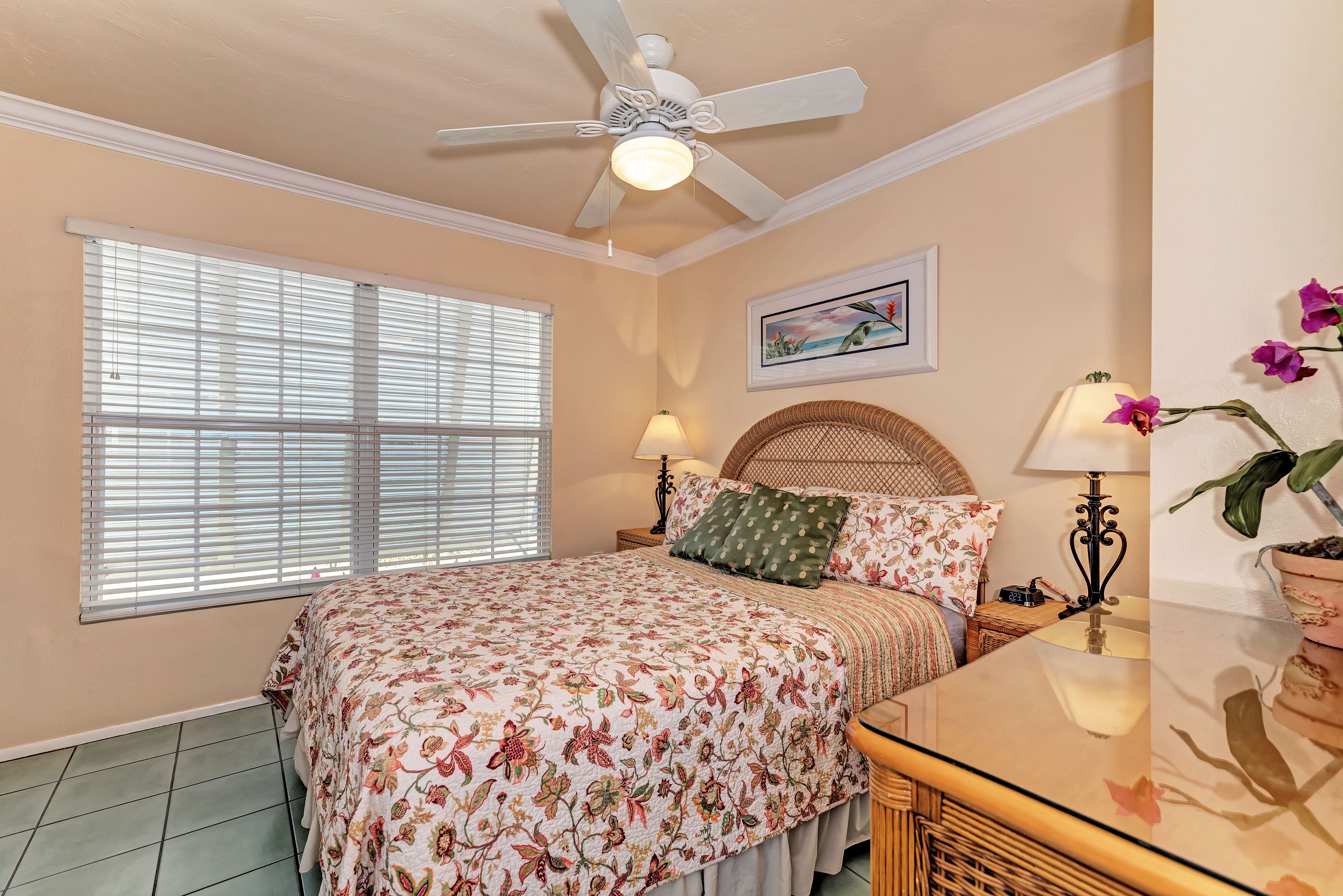 A bedroom with a bed, a ceiling fan, a window with blinds, a bedside table with a lamp, and a wall-mounted picture.