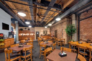 A spacious restaurant with wooden tables and chairs, a brick wall, and a ceiling with exposed pipes and lighting fixtures.