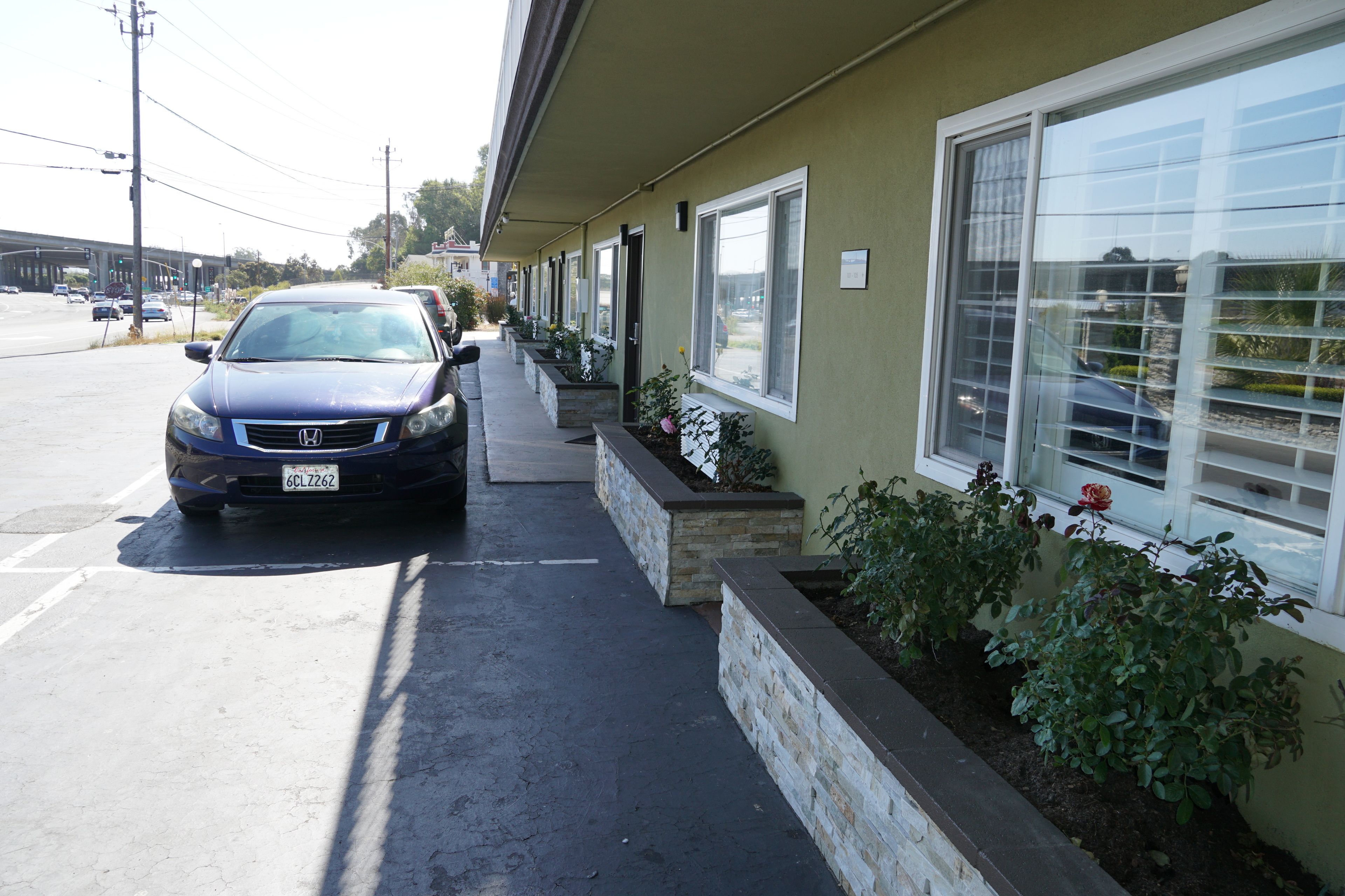 Un parking avec une voiture garée devant un immeuble doté d’une rangée de fenêtres et d’un jardinier avec des plantes.