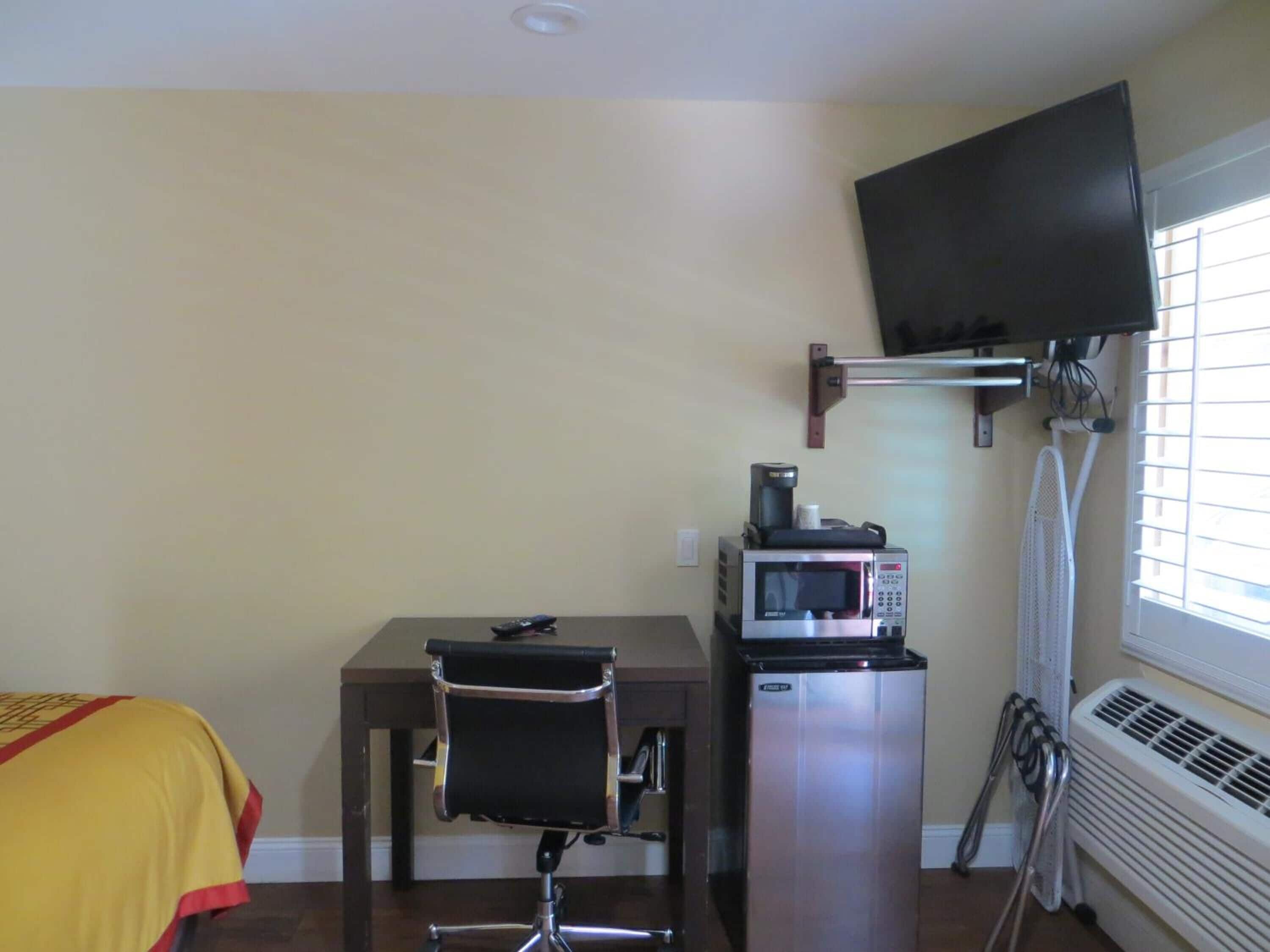 A compact room with a desk, a chair, a microwave, a small refrigerator, a television mounted on the wall, and a window with blinds.