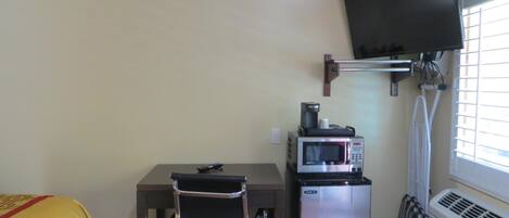 A compact room with a desk, a chair, a microwave, a small refrigerator, a television mounted on the wall, and a window with blinds.