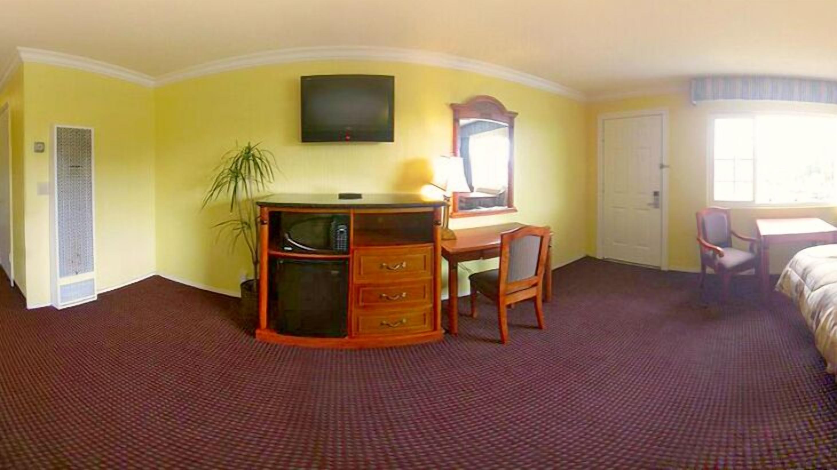 Habitación de hotel con escritorio, silla y cama. Hay un televisor montado en la pared y una ventana que proporciona luz natural.
