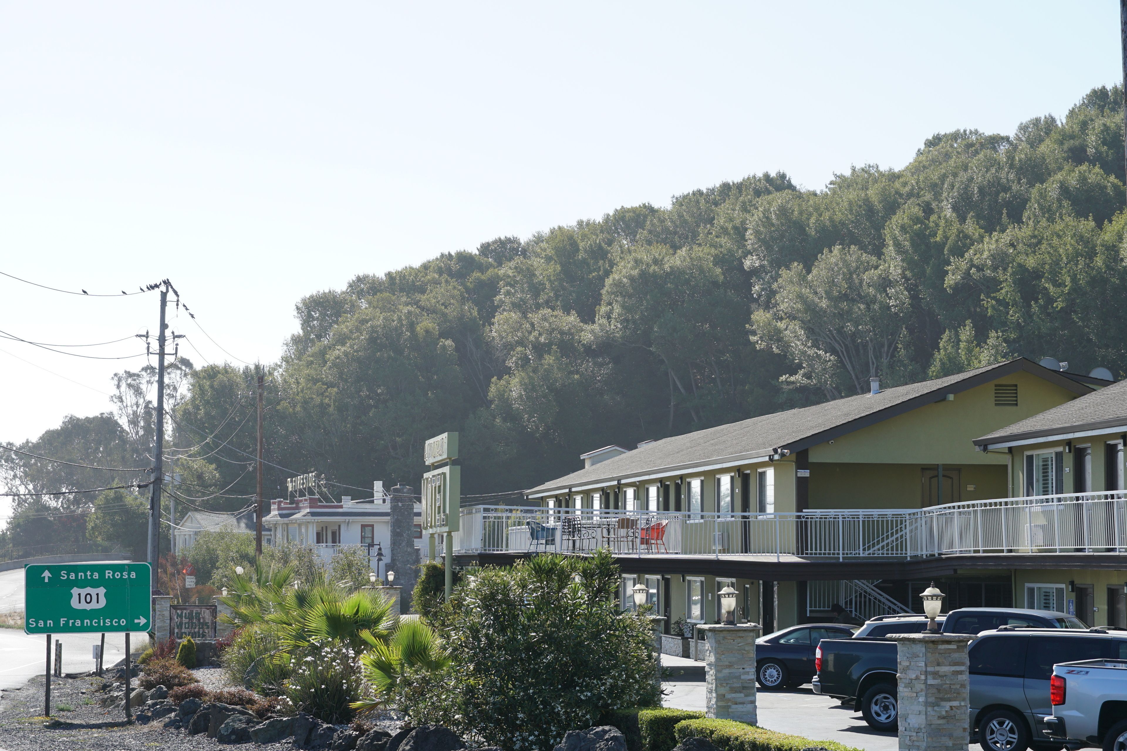 A motel with a balcony overlooking a parking area, a sign indicating Santa Rosa 101, and a backdrop of a forested hill.