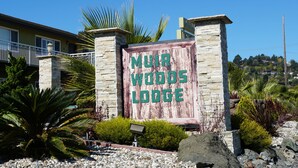 A sign for 'Muir Woods Lodge' with a stone and wooden structure, surrounded by landscaped greenery and a clear blue sky. - Muir Woods Lodge (Mill Valley)