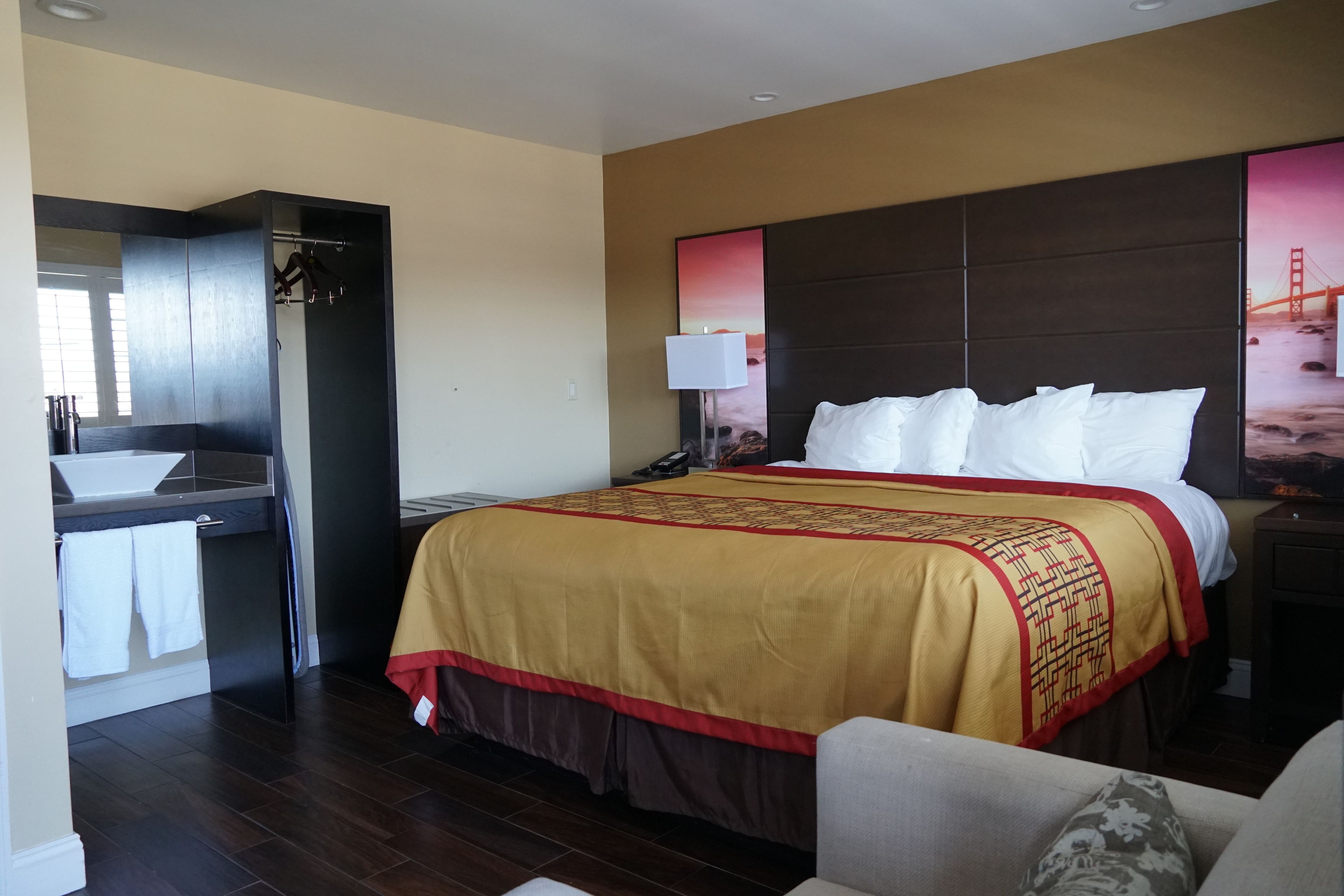 A hotel room with a large bed, a bedside table, a nightstand, a mirror, a wardrobe, and a bathroom with a sink and towel rack.