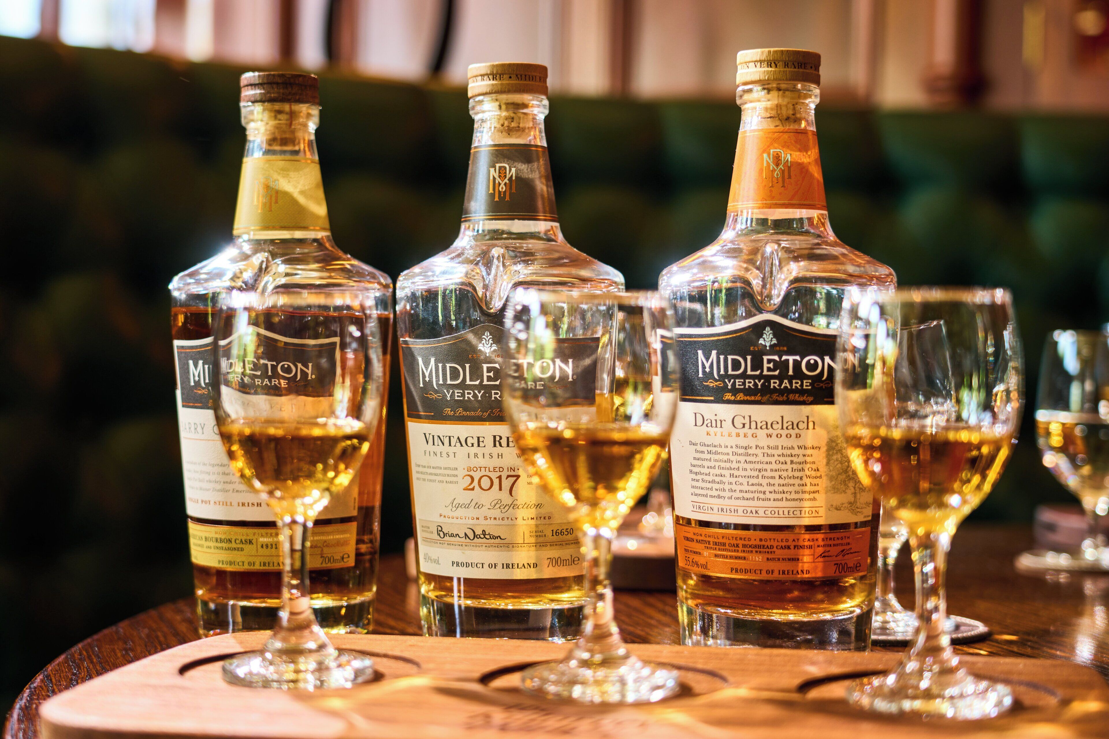 Drie flessen Midleton Very Rare Ierse Whiskey en twee glaasjes op een houten oppervlak.