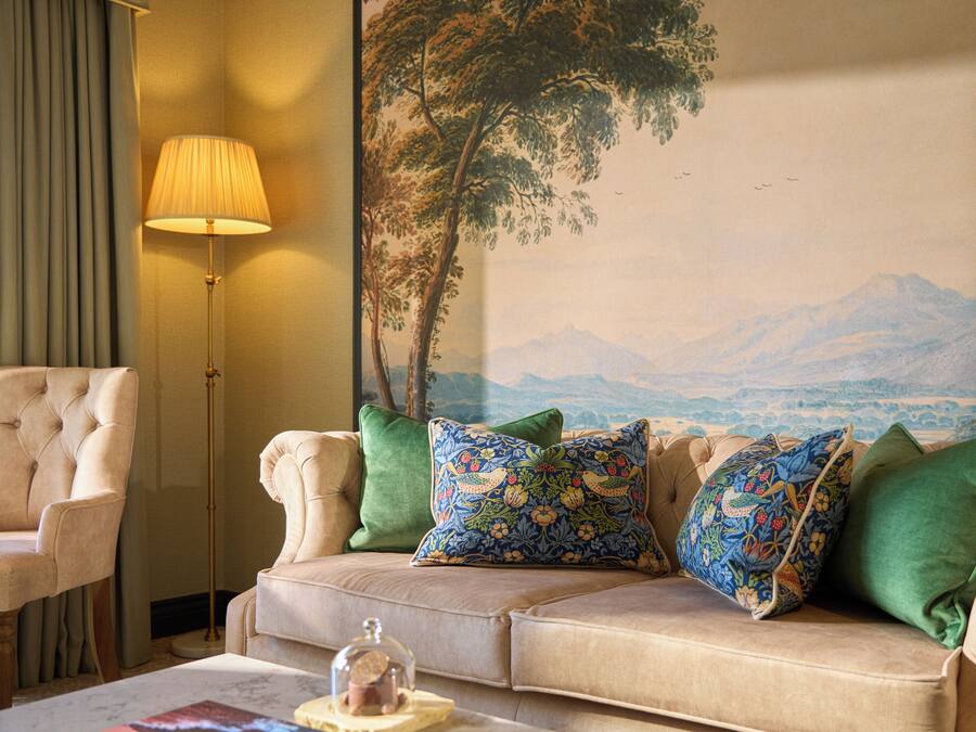 Una sala de estar con un sofá color beige, almohadones estampados en verde y azul, una lámpara de pie y un mural grande con un paisaje montañoso.