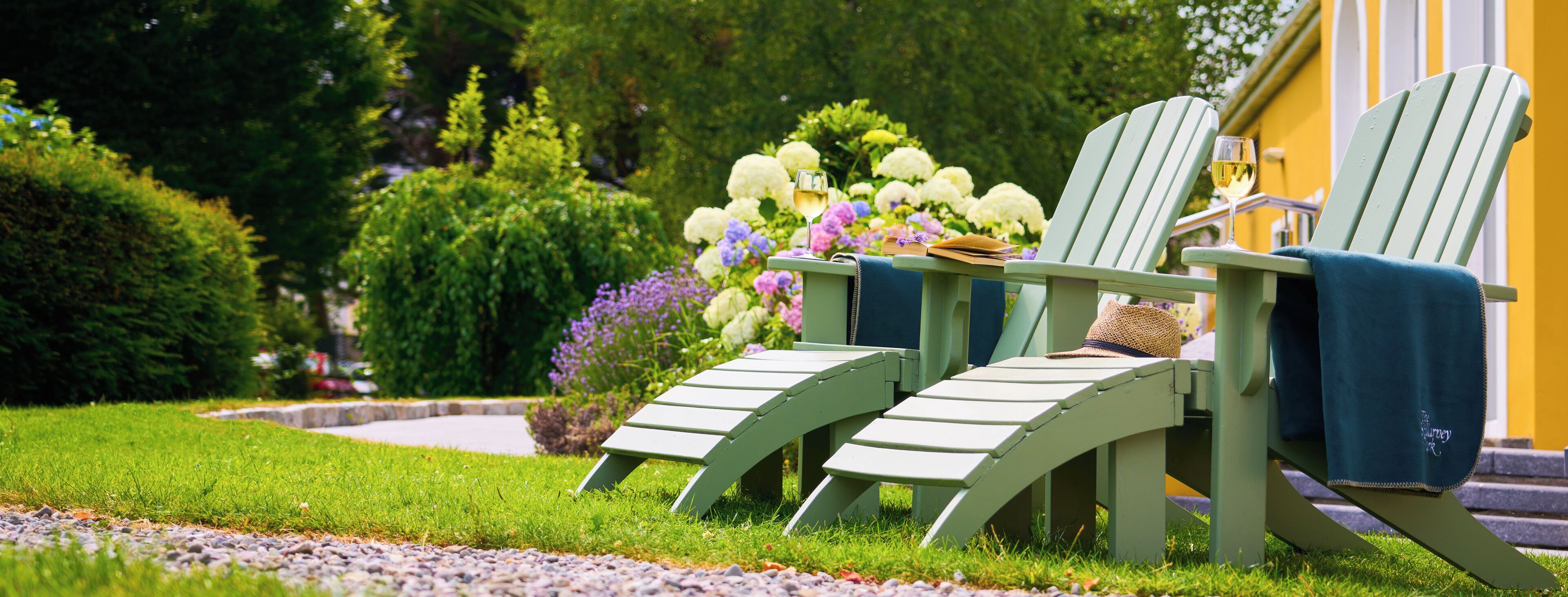 Two green Adirondack chairs with a glass of white wine on a garden bench, surrounded by lush greenery and flowers.