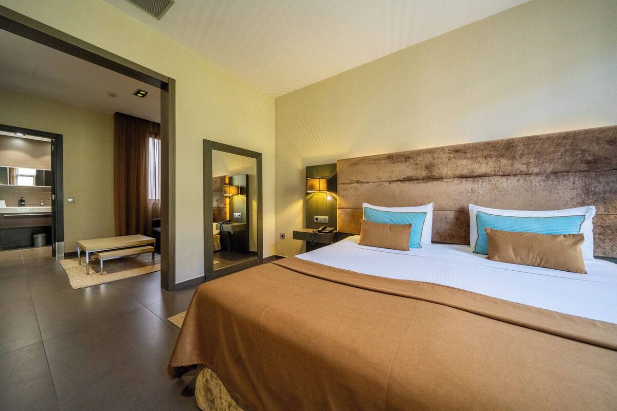 Una habitación de hotel con una cama grande, una mesita de noche, un tocador, un espejo y un baño visible a través de una puerta abierta.