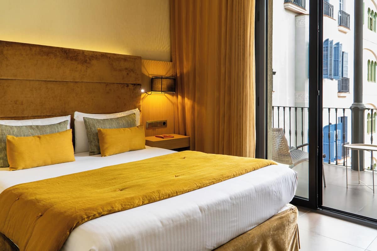 Habitación de hotel con una cama grande, una mesita de noche, una lámpara y vistas al balcón exterior.