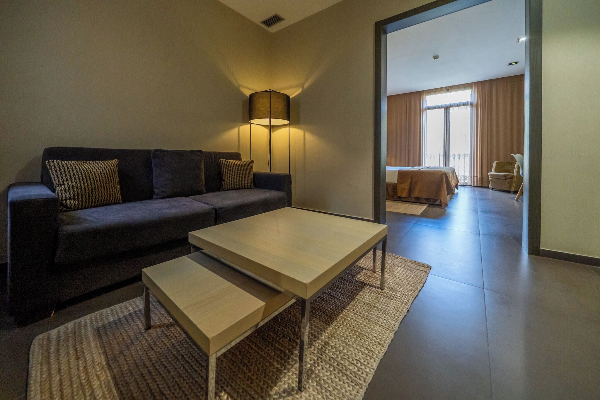 A modern living room with a sofa, a coffee table, and a lamp. There is a view of a balcony with a bed and a chair.