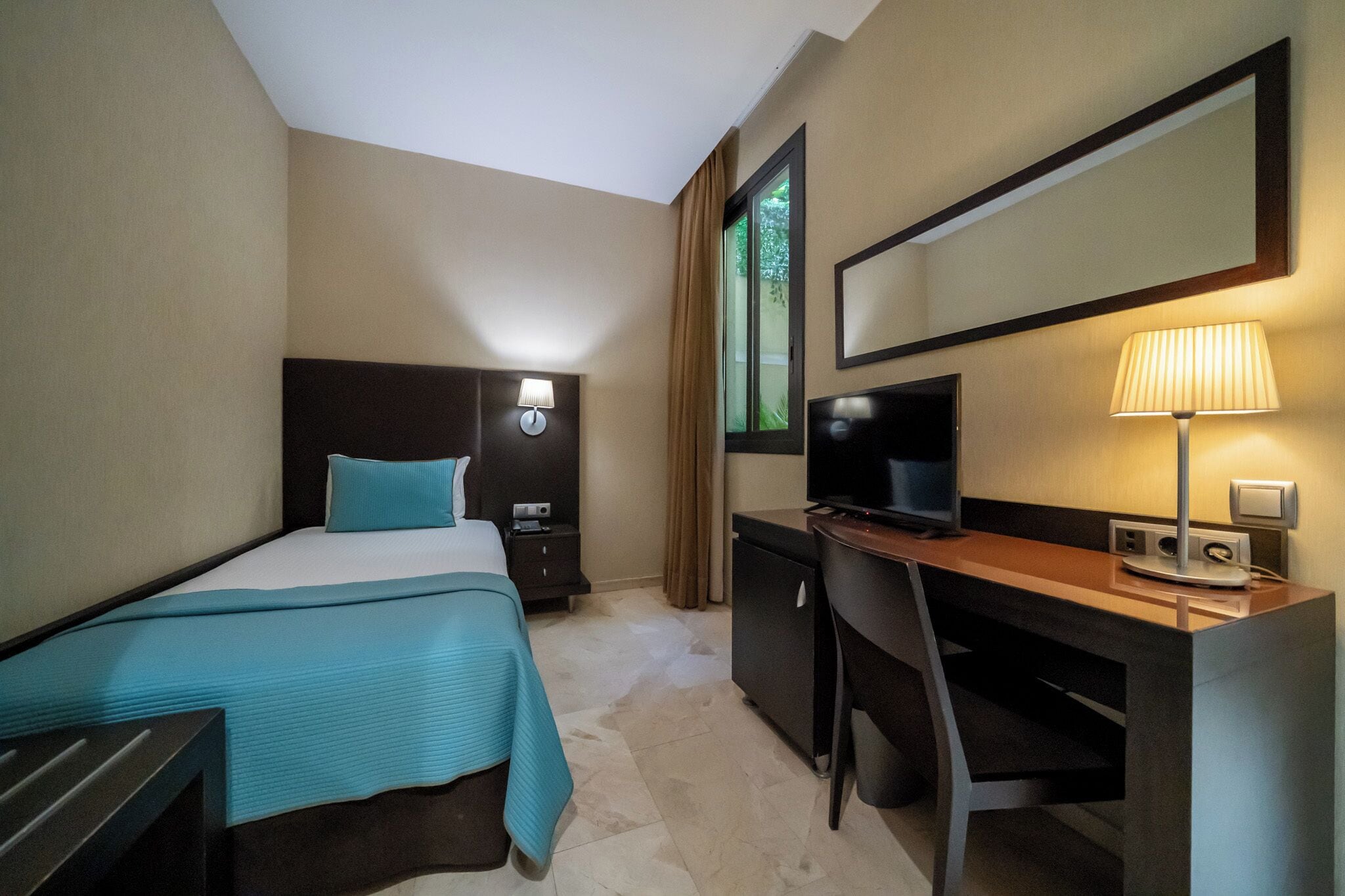 A hotel room with a bed, desk, chair, TV, lamp, and a mirror.