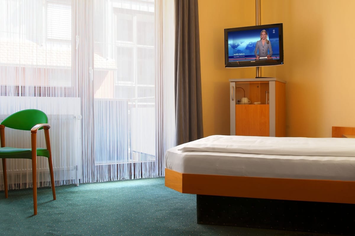 A hotel room with a bed, a television on a stand, a green chair, and a window with sheer curtains.