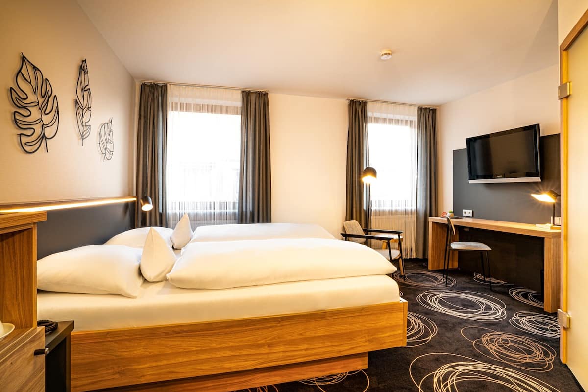 A hotel room with a double bed, a desk with a chair, a television, and a window with curtains.