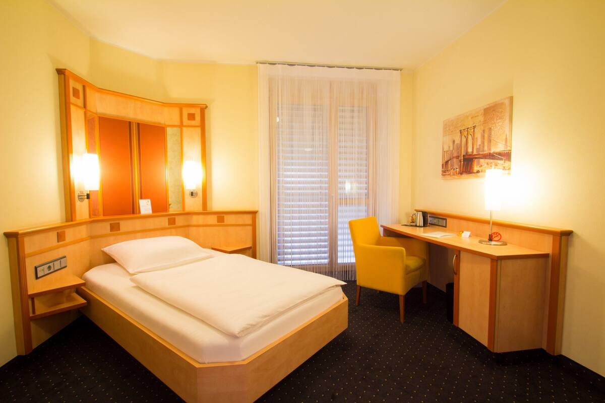 A hotel room with a bed, desk, chair, and a large window with blinds.