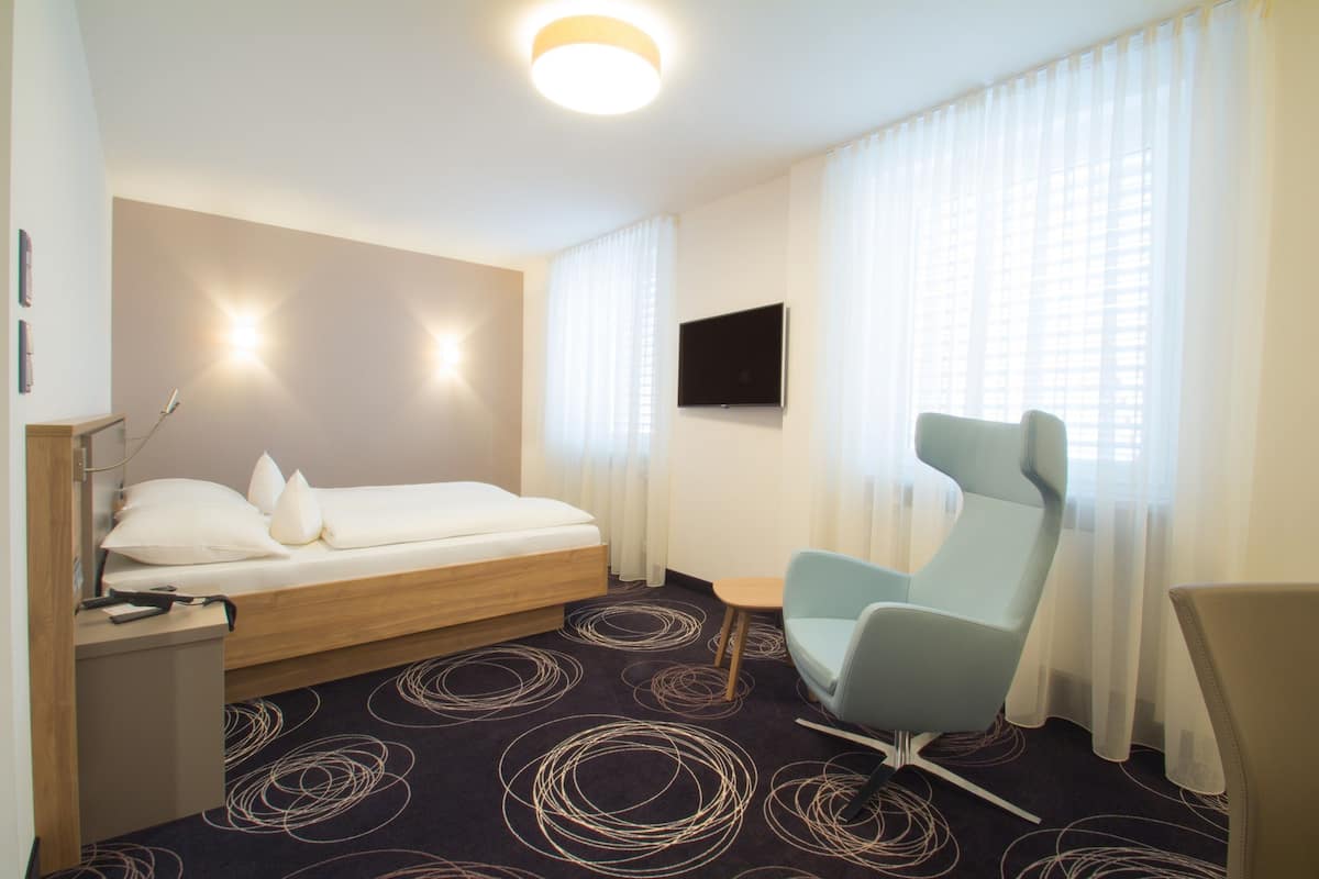 A hotel room with a bed, a desk, a chair, a TV, and a window with curtains.