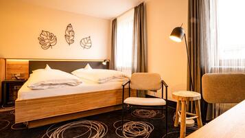 Una habitación de hotel con cama, una silla, un taburete pequeño, un escritorio y una lámpara.