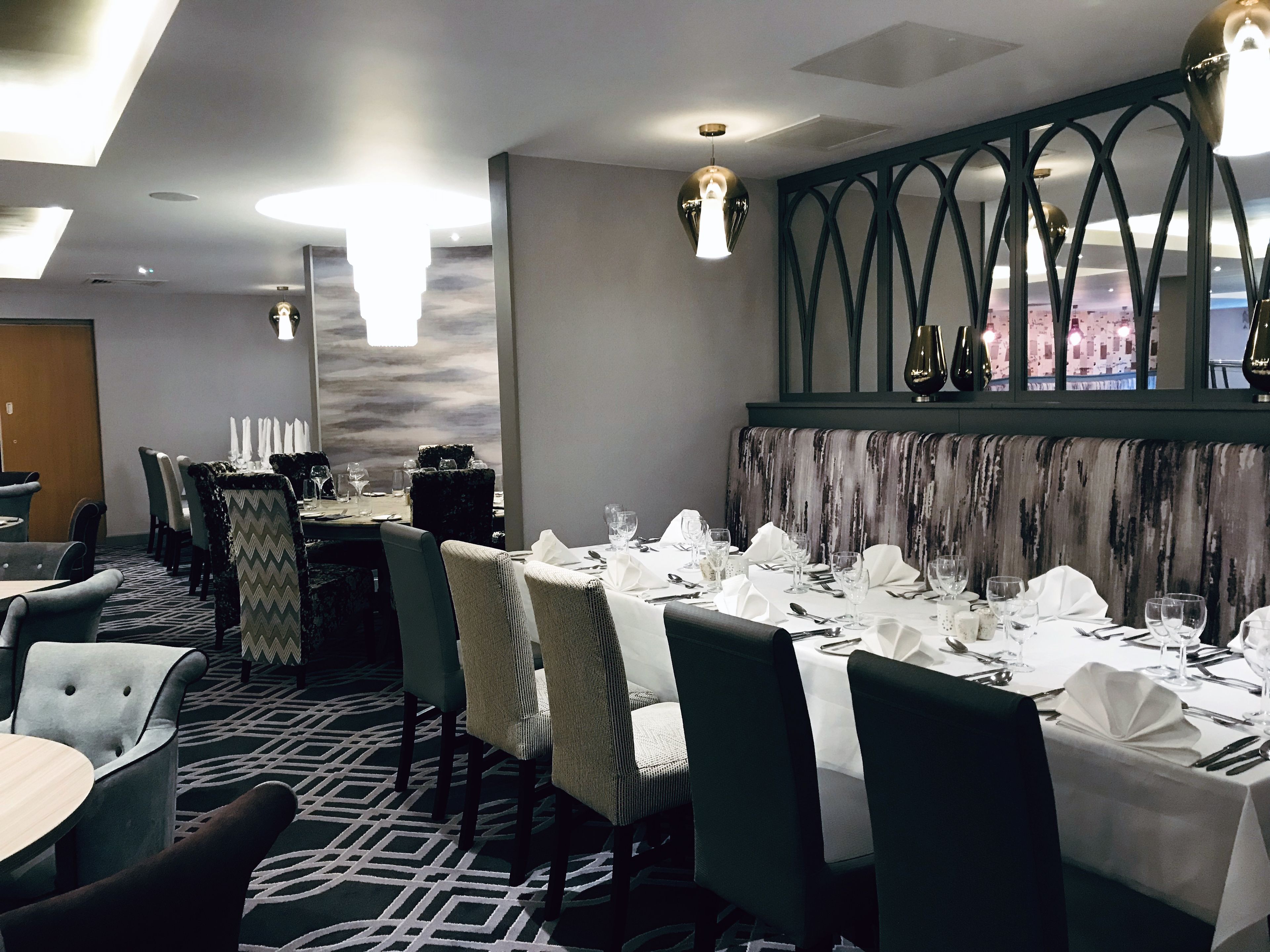 A dining area with round tables set for four, each with white tablecloths and silverware. The chairs are upholstered in a light fabric, and the carpet has a geometric pattern.