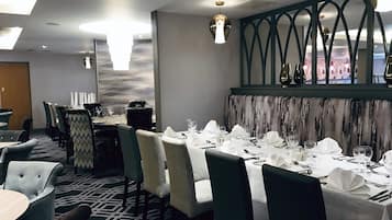 A dining area with round tables set for four, each with white tablecloths and silverware. The chairs are upholstered in a light fabric, and the carpet has a geometric pattern.