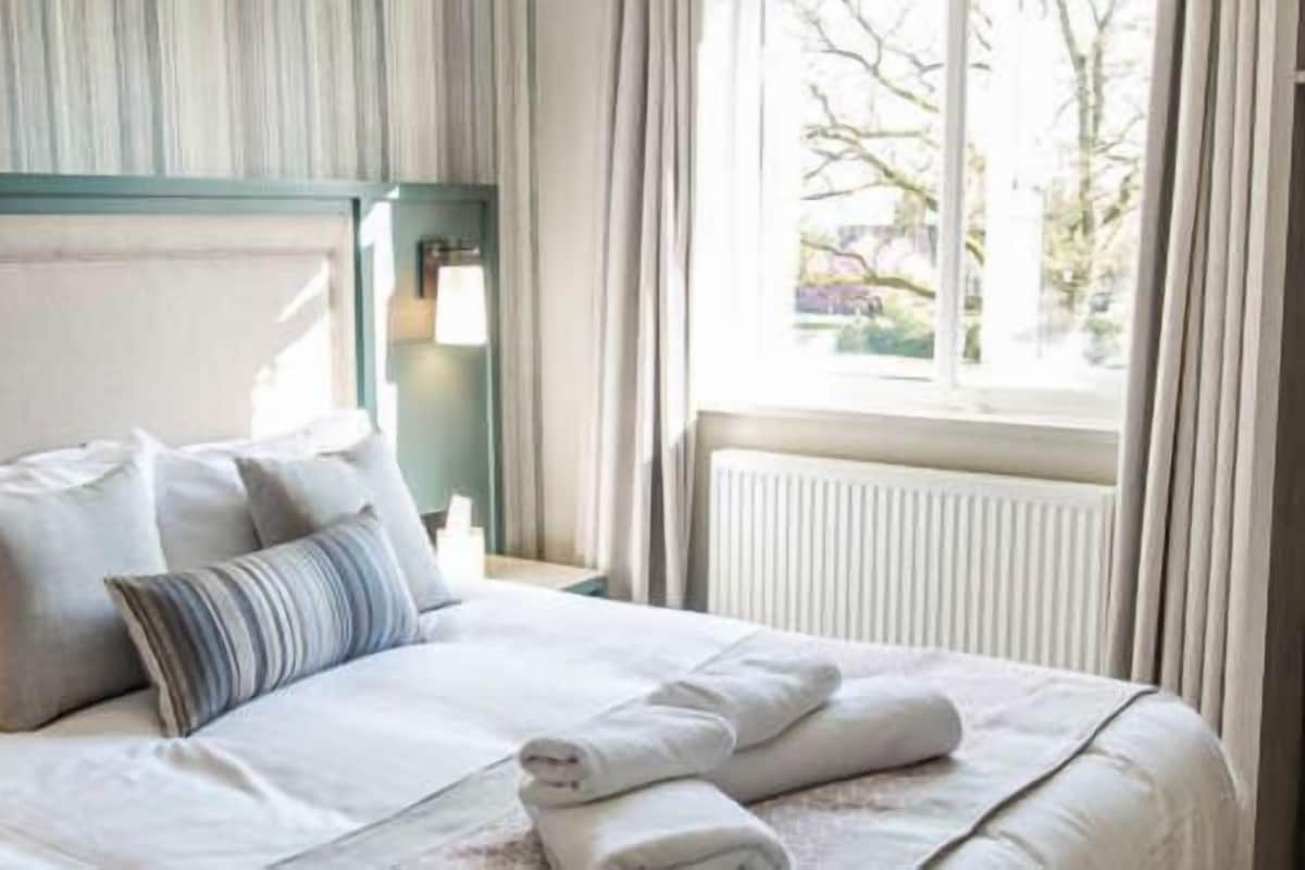 A neatly made bed with pillows and a rolled towel, a bedside table with a lamp, and a window with curtains.