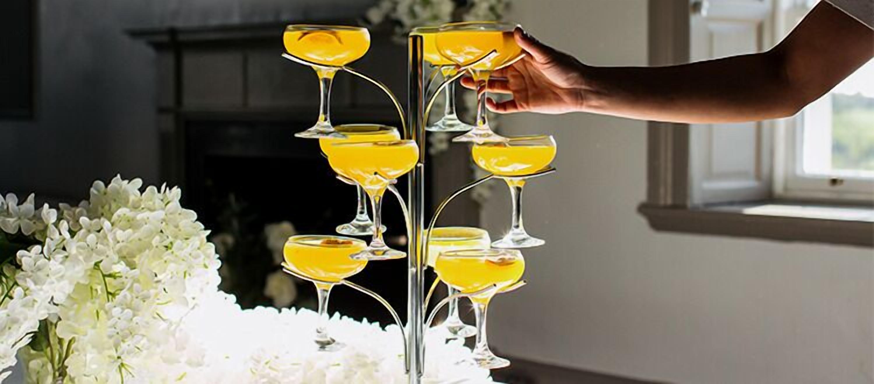 A hand holding a glass of yellow liquid, with multiple glasses arranged in a pyramid on a table, surrounded by white flowers.